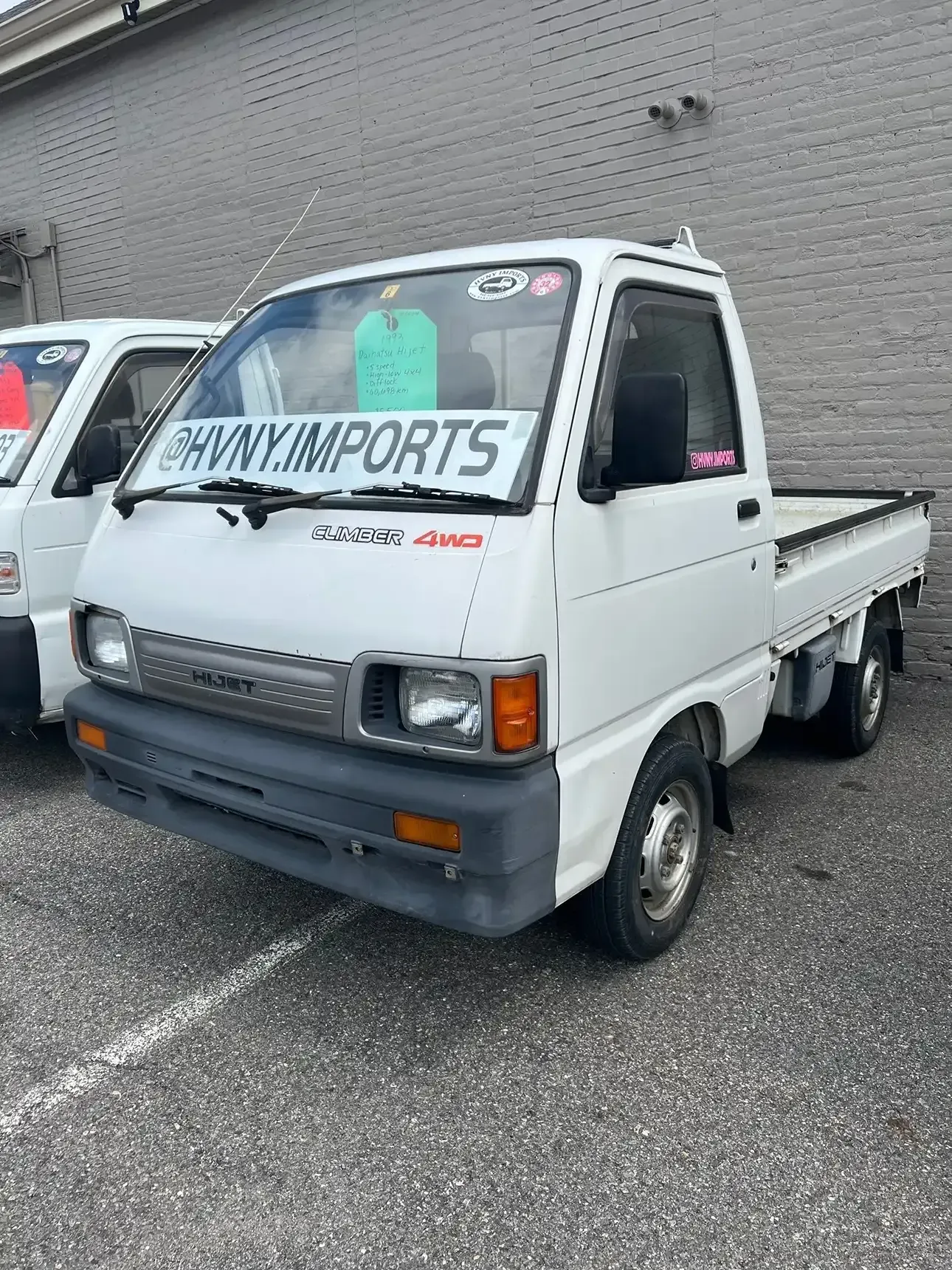 A white Daihatsu Hijet kei truck parked on pavement next to another vehicle with a large HVNY.IMPORTS decal on the glass.