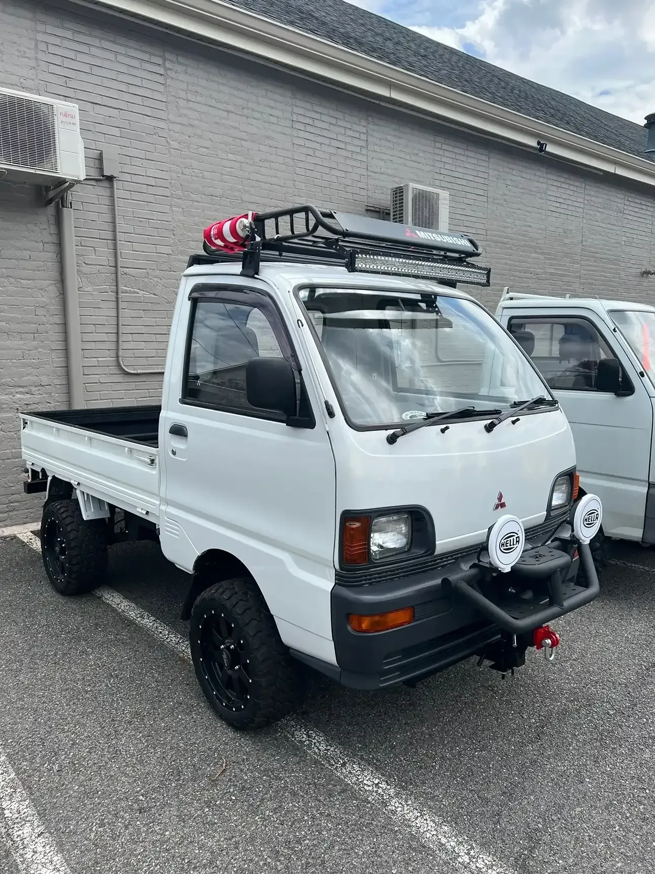 A white, lifted Mitsubishi mini truck with a roof rack and off-road lights, parked in a parking lot.