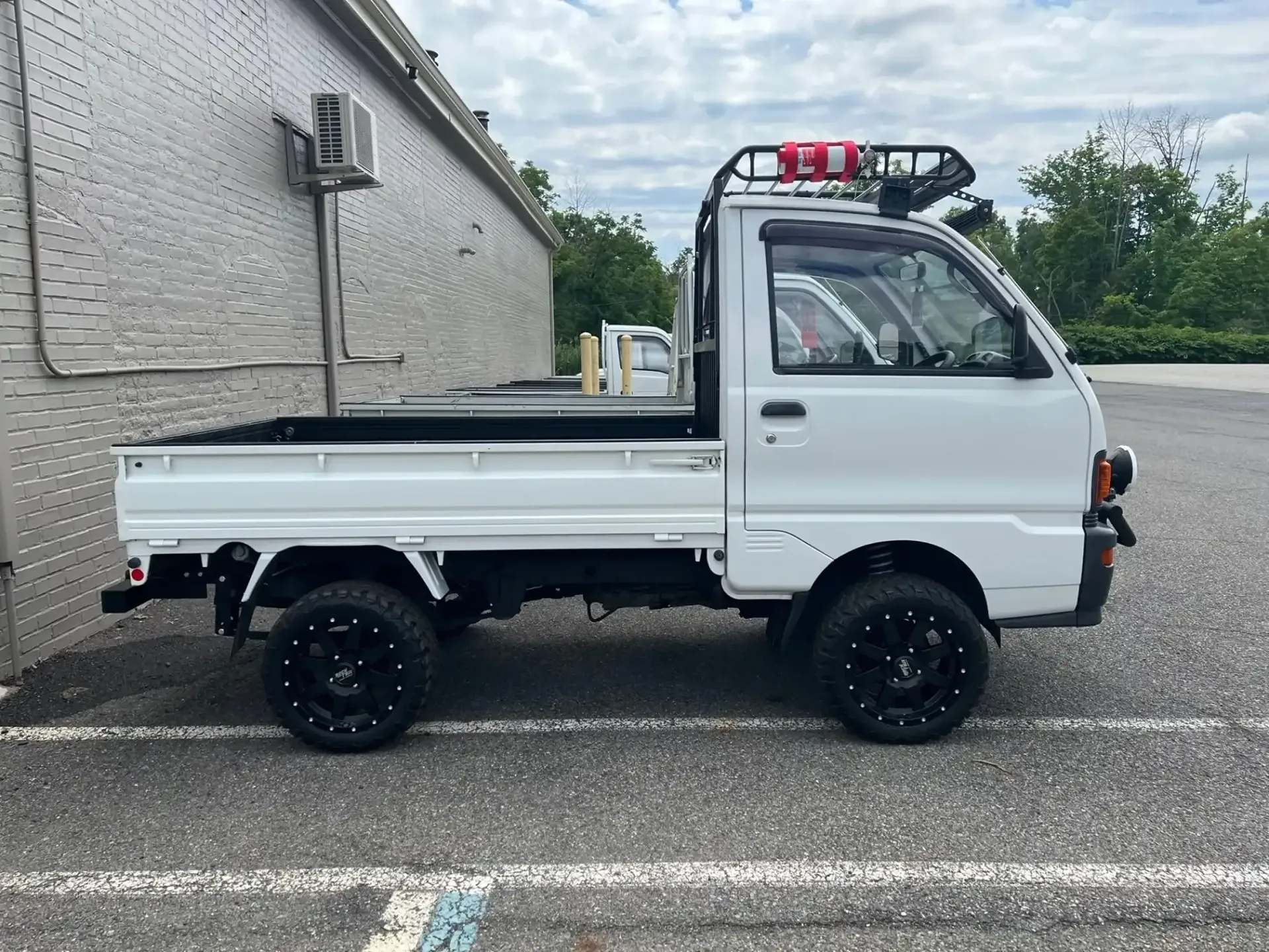 A white, lifted Kei truck with black aftermarket wheels and a roof rack, parked on an asphalt lot beside a brick wall.