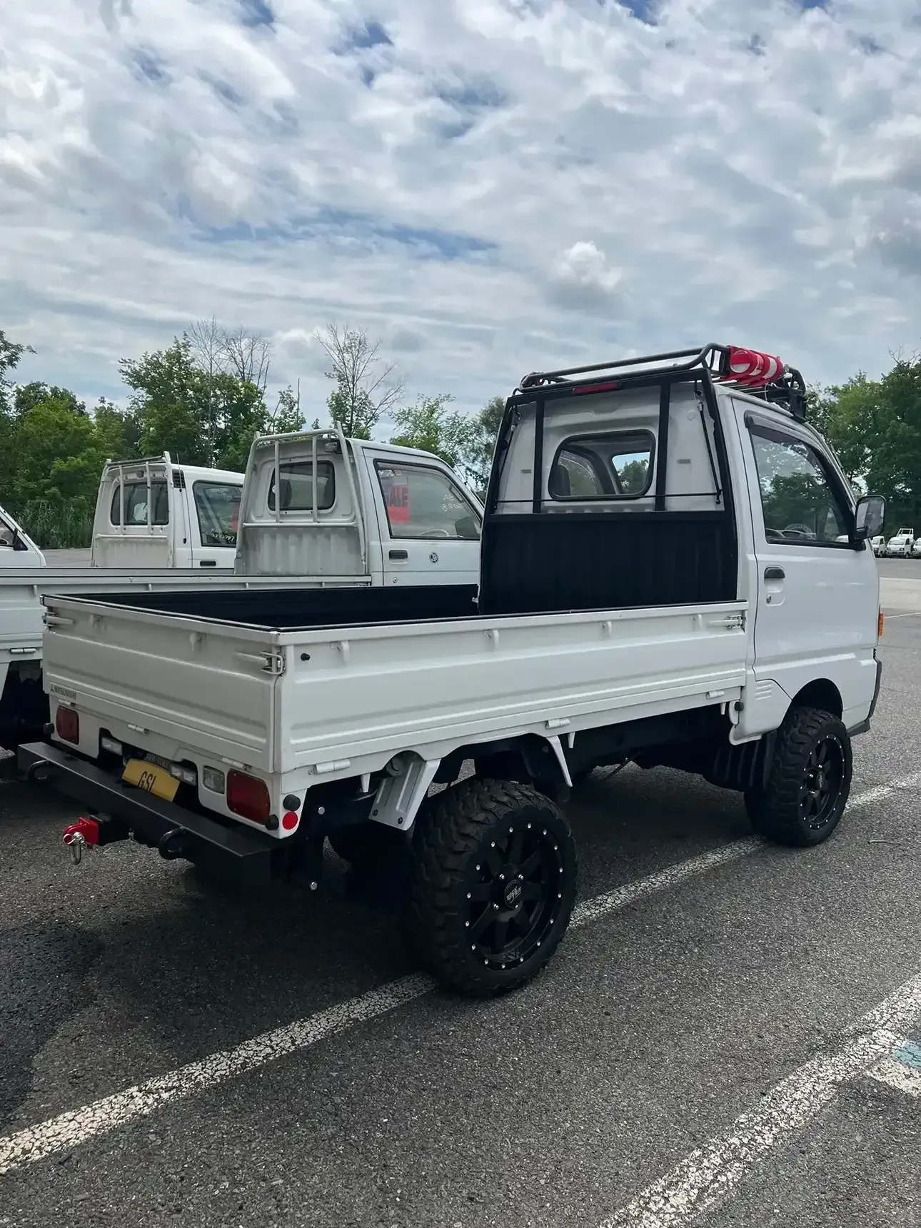 A white Kei truck with off-road tires and a roof rack parked in an outdoor lot.