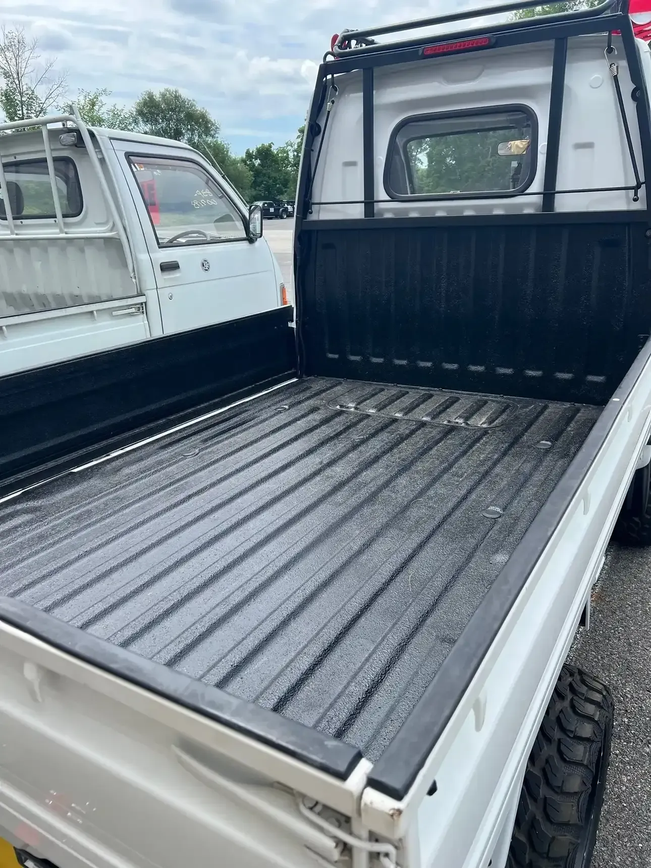 White mini trucks parked outdoors, focusing on the empty black cargo bed and protective metal rack of the foreground one.