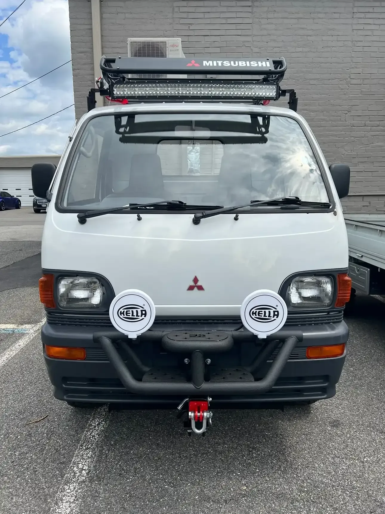 Front view of a white Mitsubishi Kei truck with a roof rack, light bar, off-road bumper, and two large Hella lights.