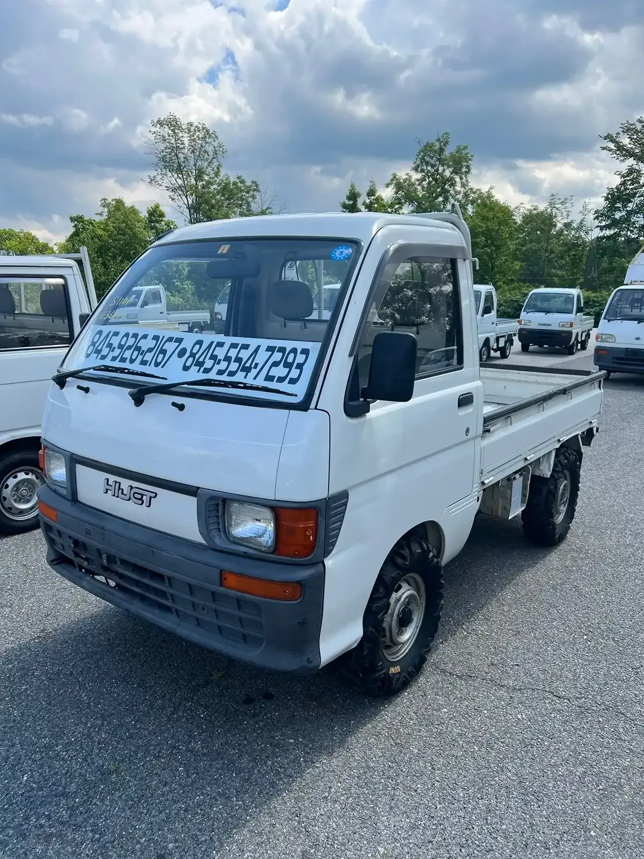 A white Mitsubishi Minicab kei truck parked outdoors with phone numbers displayed on the windshield.