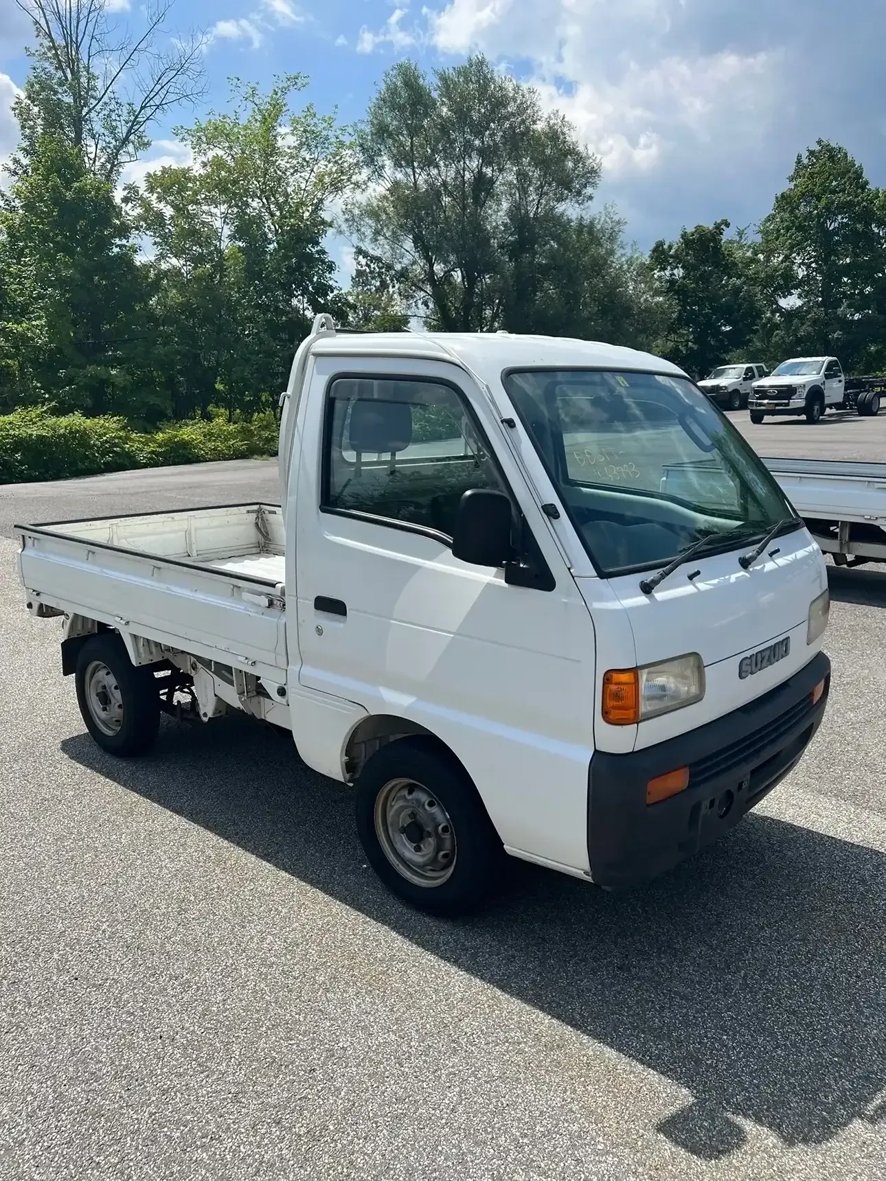A white kei truck parked on an asphalt lot with trees and a cloudy sky in the background.