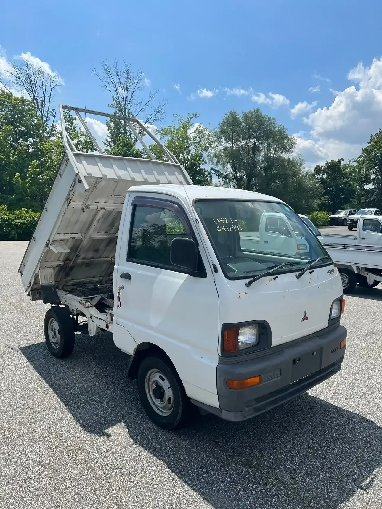 A white Mitsubishi mini-dump truck parked on a gravel lot under a clear blue sky, with its cargo bed in a raised position.