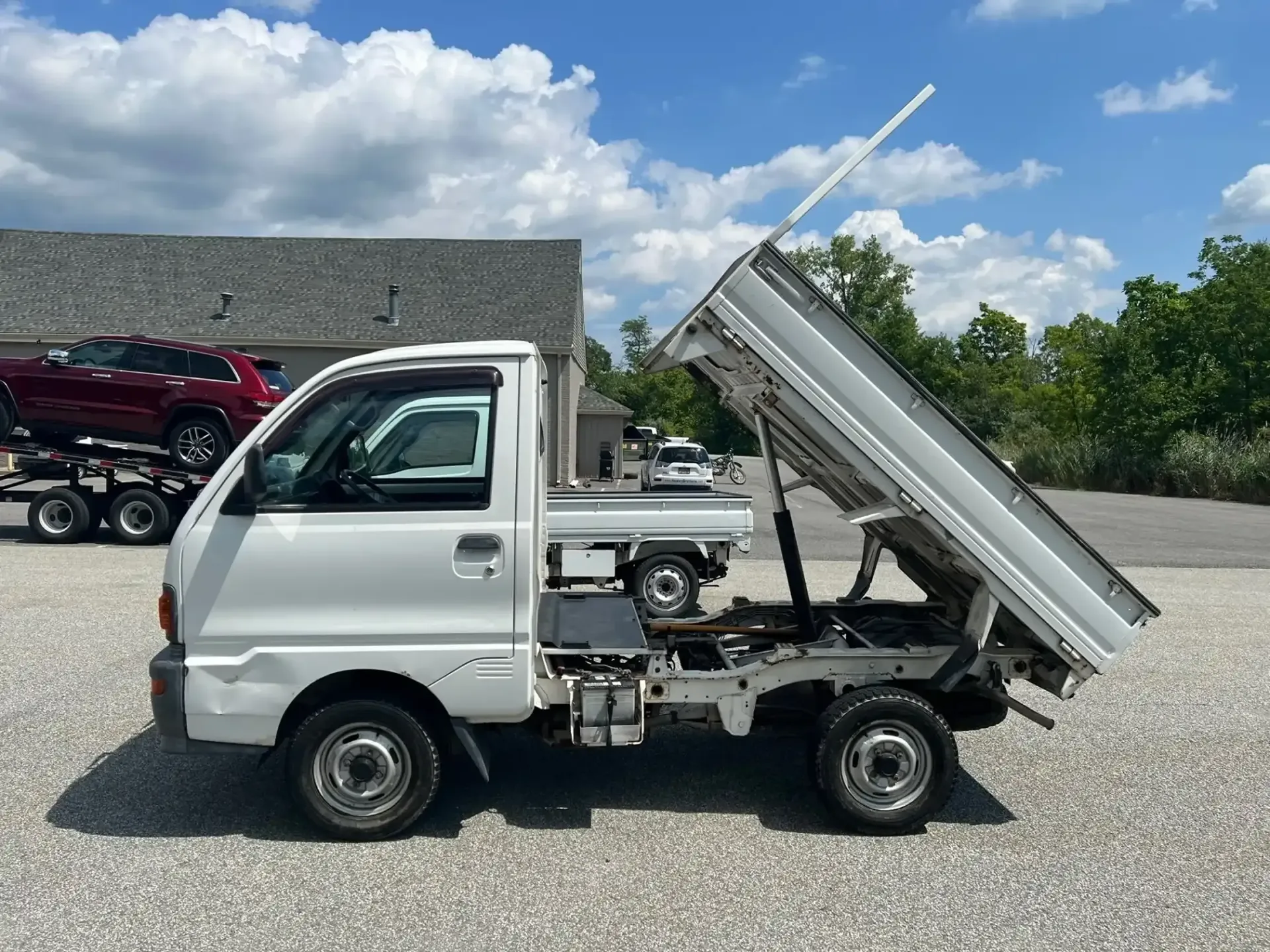A white kei dump truck sits on a gravel lot under a sunny sky with its cargo bed tilted upward in the dumping position.