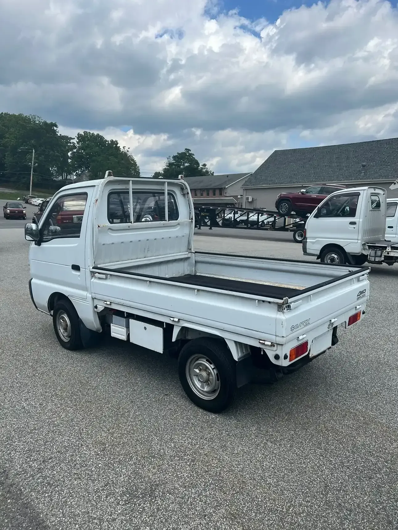 A white kei truck parked on a gravel lot under a cloudy sky, seen from a rear three-quarter angle.