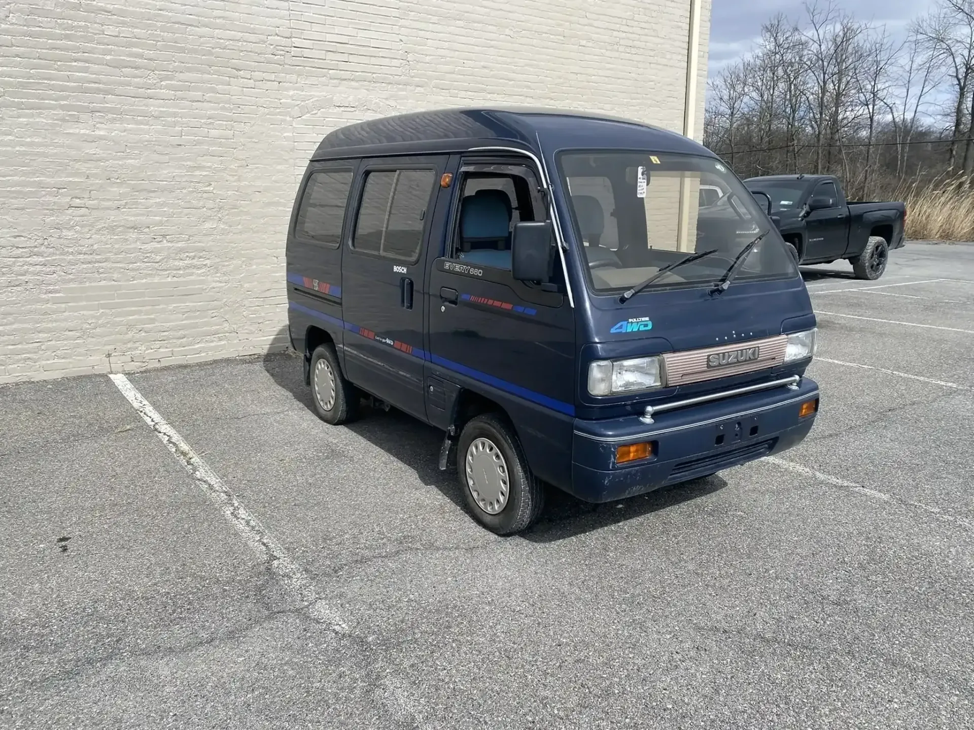 A small, blue microvan parked in a gravel lot next to a light-colored brick wall.