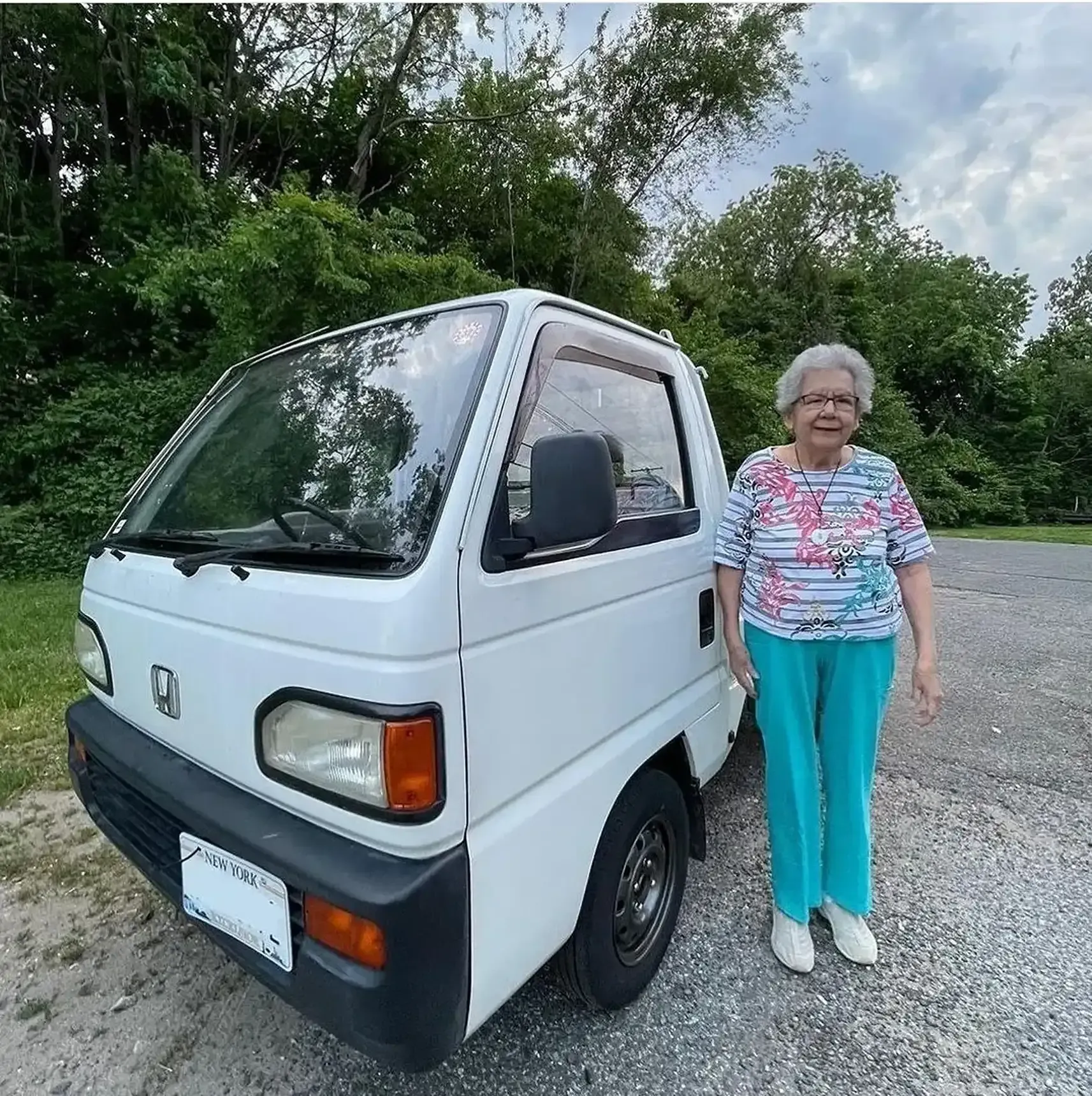 An elderly person stands smiling beside a small, white Honda kei truck parked on a gravel path near green trees.