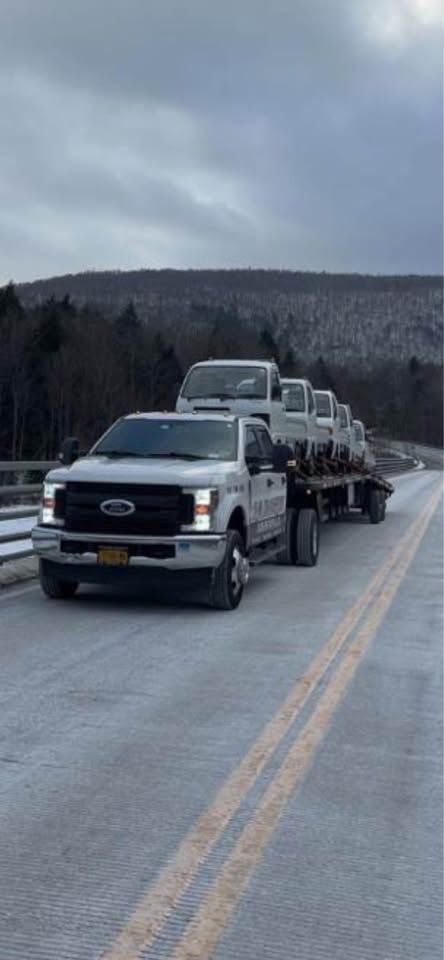 A white Ford pickup truck towing a flatbed trailer loaded with several white, compact utility vehicles on a snowy road.