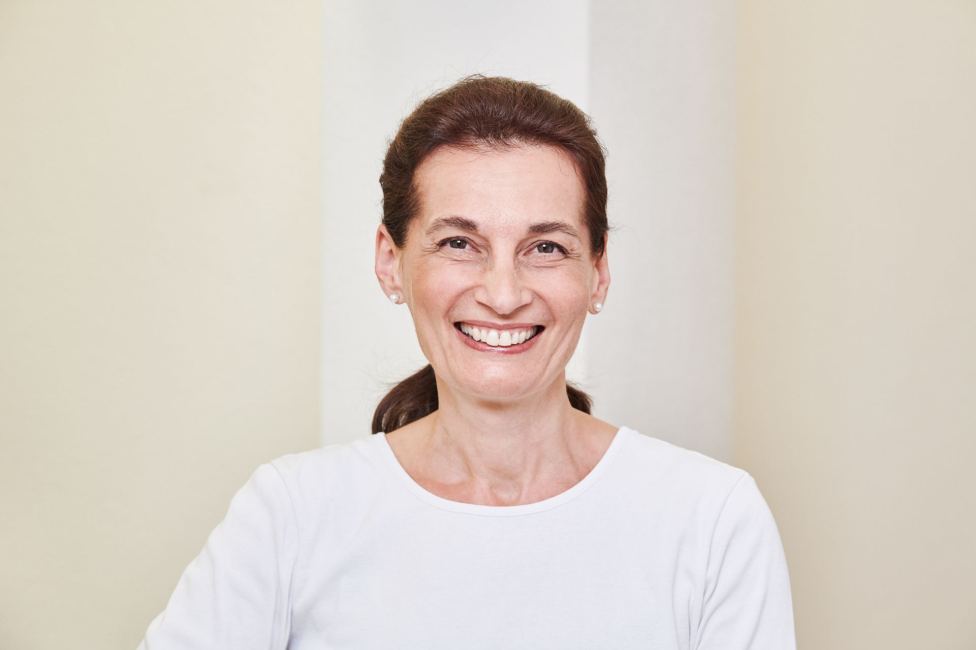 Eine lächelnde Frau mit zurückgebundenem, dunklem Haar, die ein weisses Hemd und eine Perlenkette trägt.