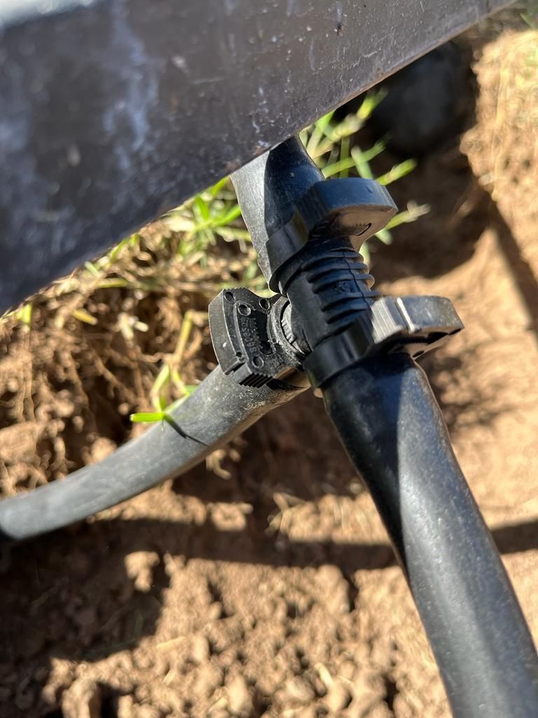Tubo de irrigación negro conectado a un codo de plástico negro, colocado en tierra cerca de un borde gris.