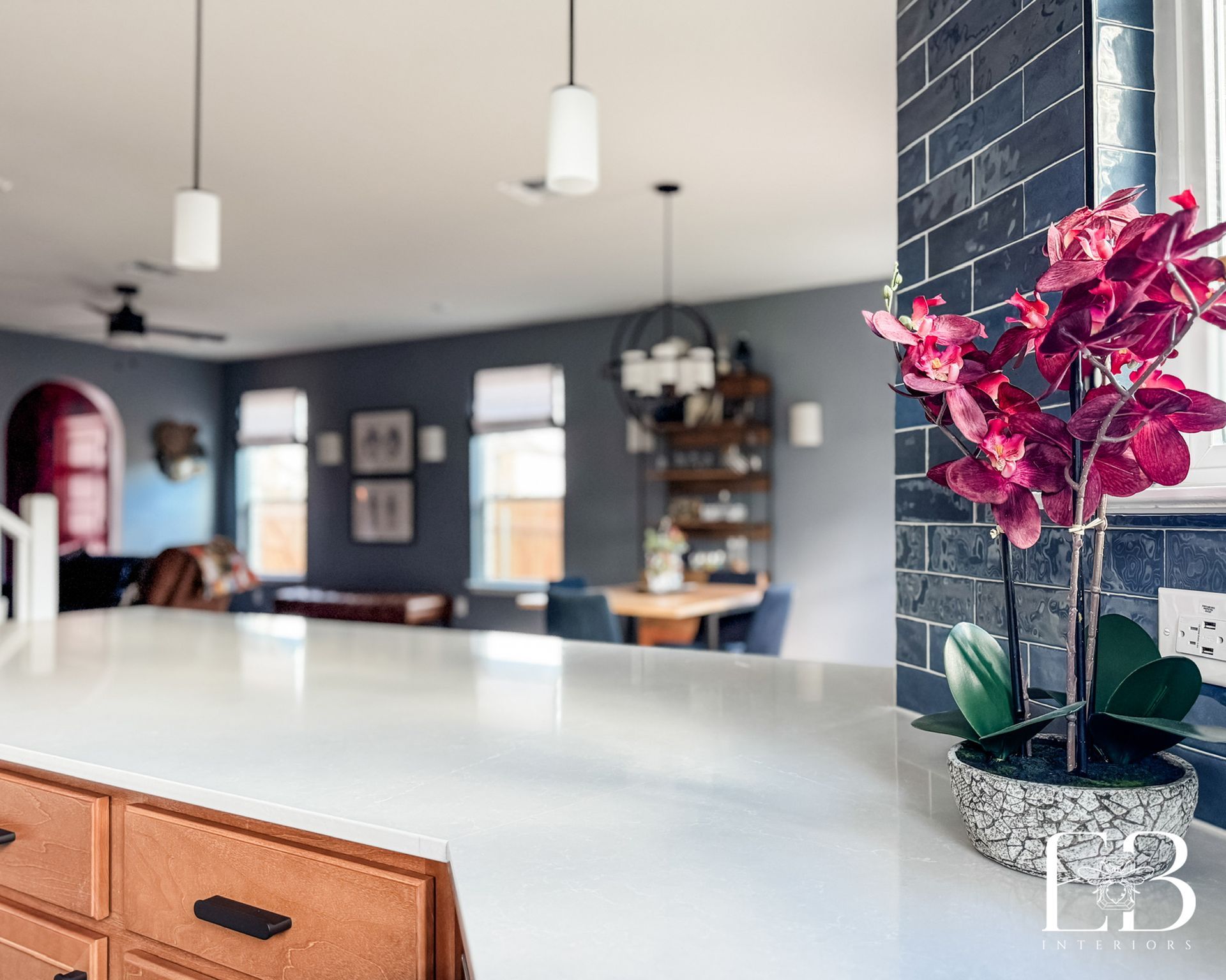Dining area with wood table, navy chairs, and chandelier overlooking a blue-tiled kitchen by Emerald Bee Interiors.