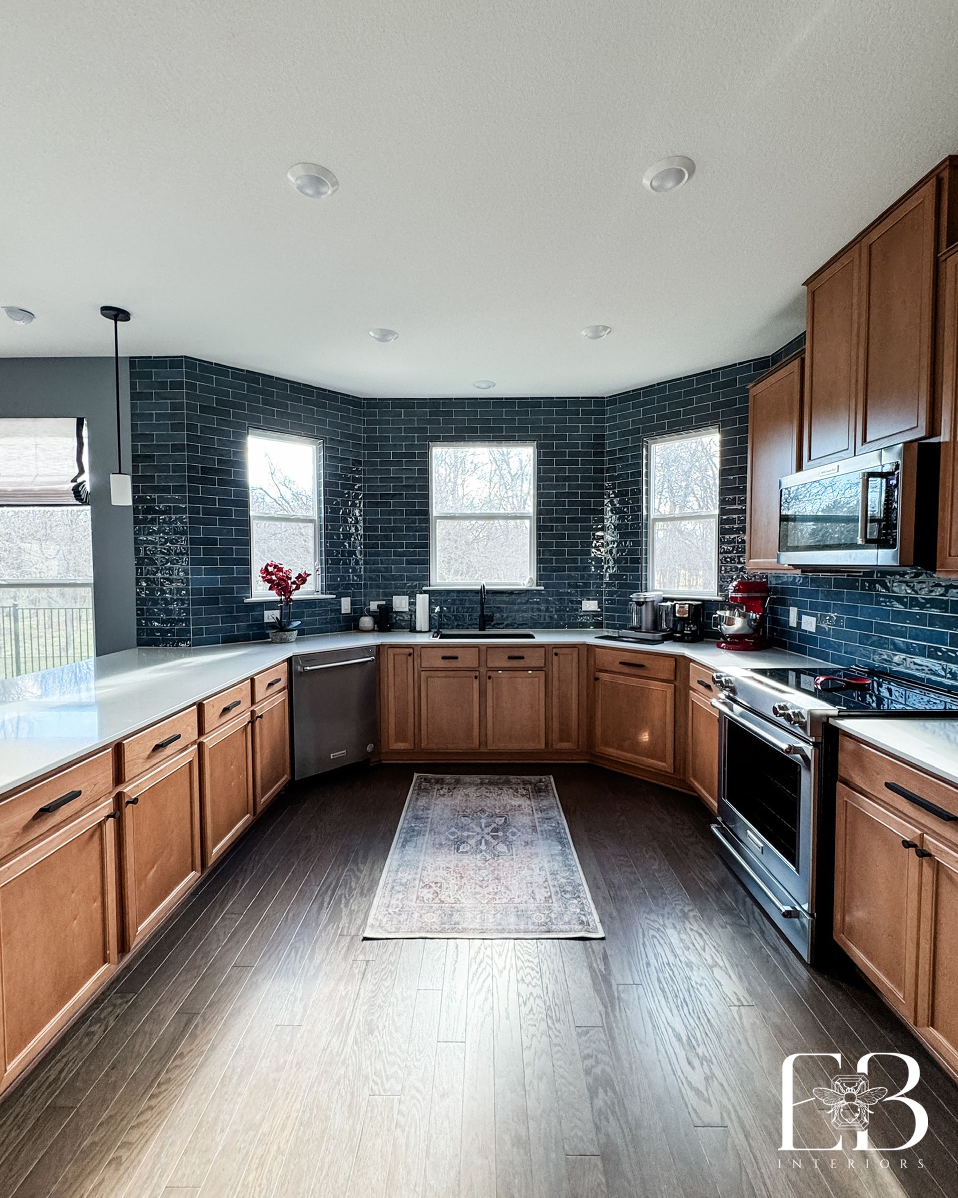 Modern kitchen by Emerald Bee Interiors with warm wood cabinetry, blue tile backsplash, and large windows for natural light.
