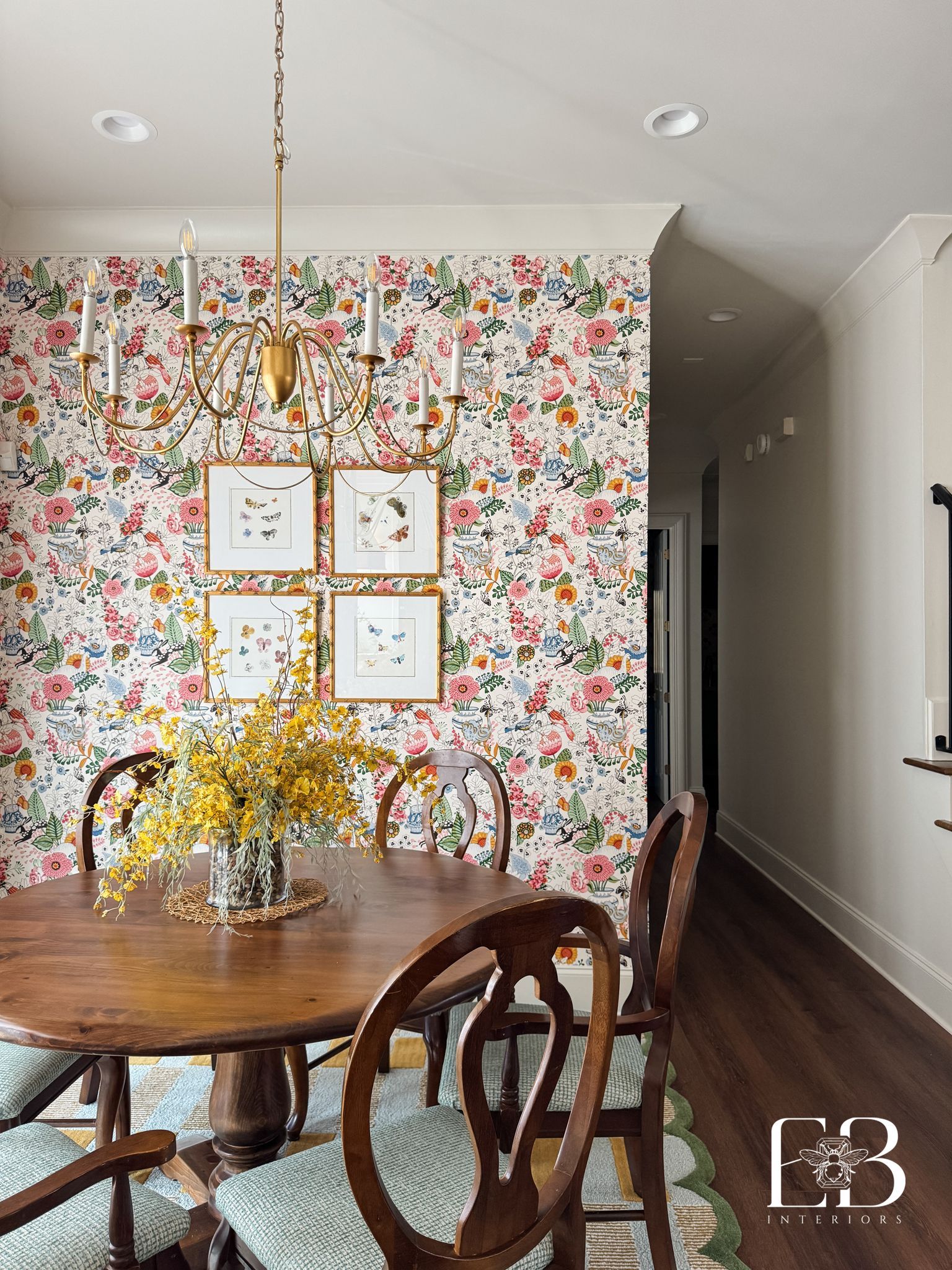 Dining room with floral wallpaper, round wooden table, chairs, and gold chandelier.