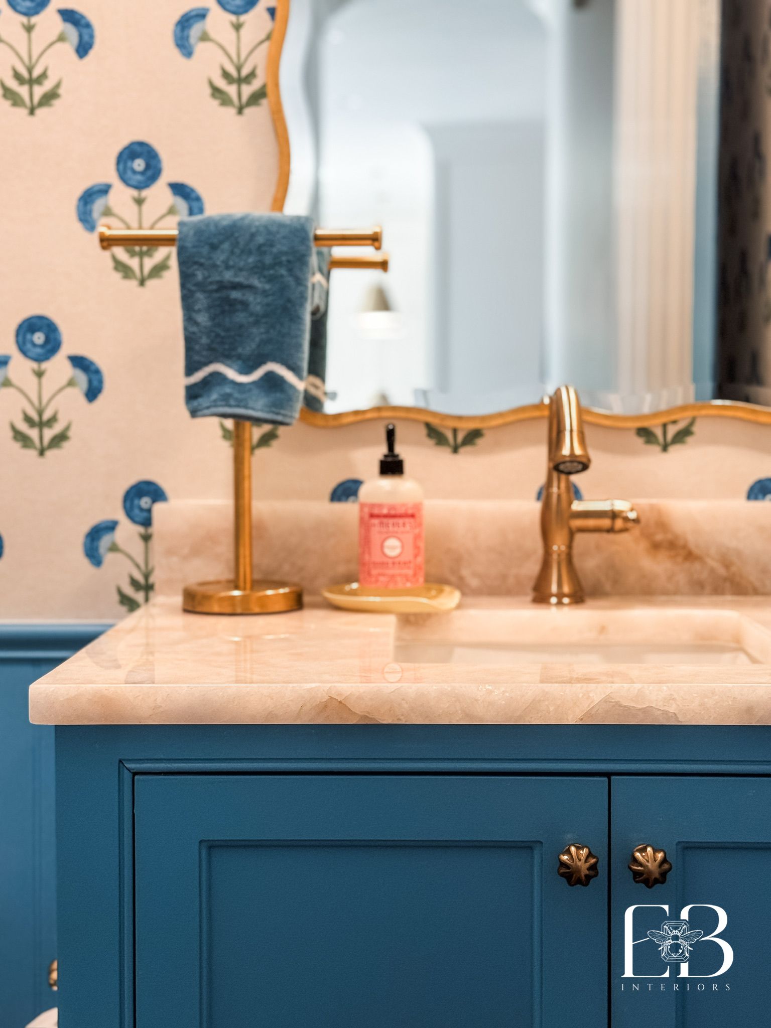 Powder bath with blue vanity, brass fixtures, and floral Schumacher wallpaper by Emerald Bee Interiors.