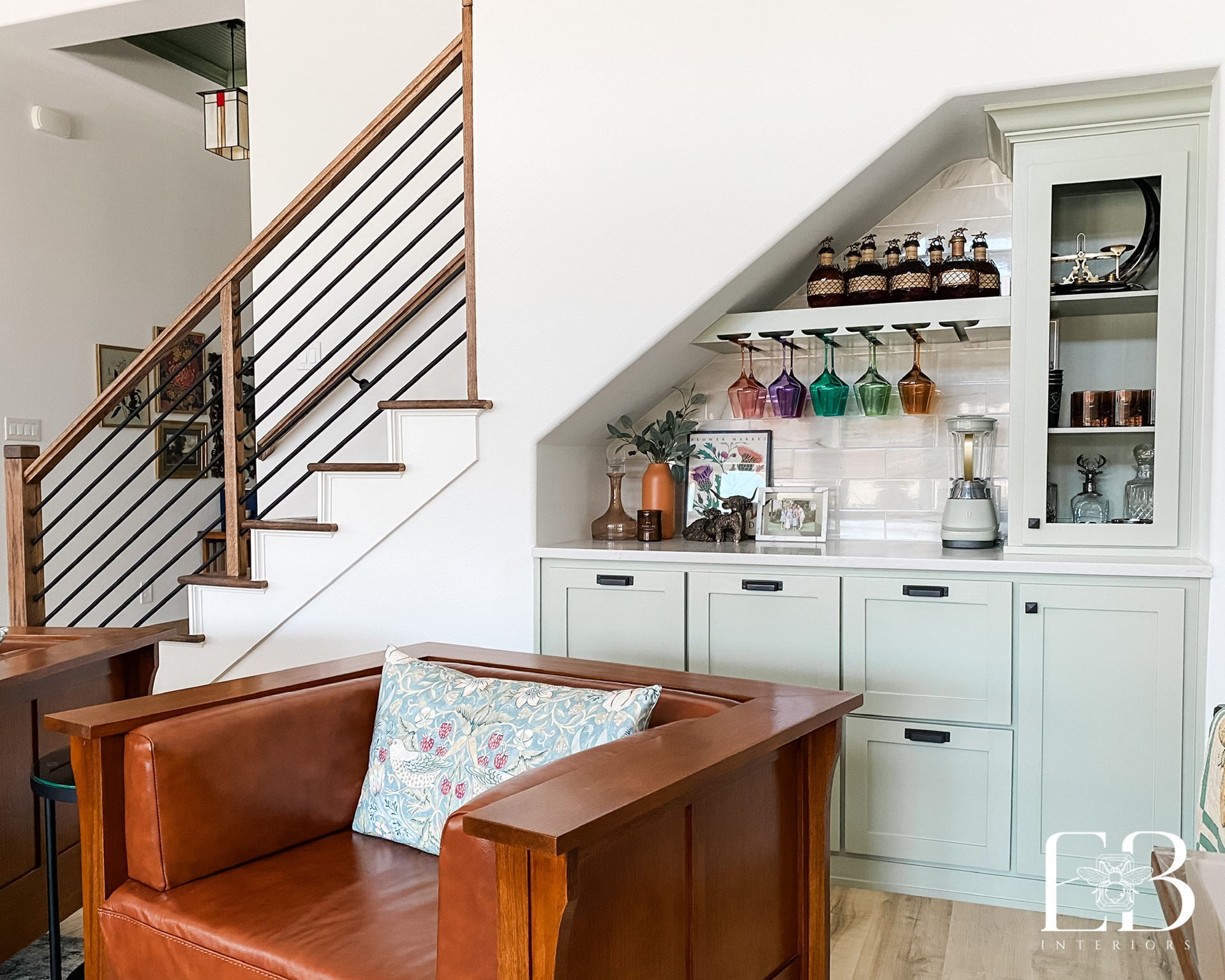 Built-in bar under staircase by Emerald Bee Interiors with sage cabinetry, black hardware, and rainbow glassware display.