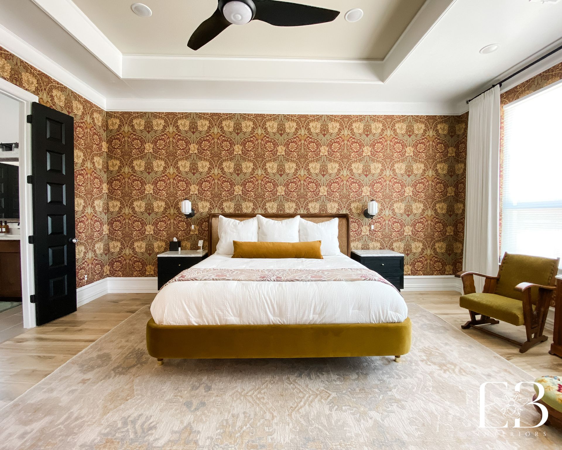 Primary suite with gold and red floral wallpaper, layered textures, and a mustard upholstered bed by Emerald Bee Interiors.