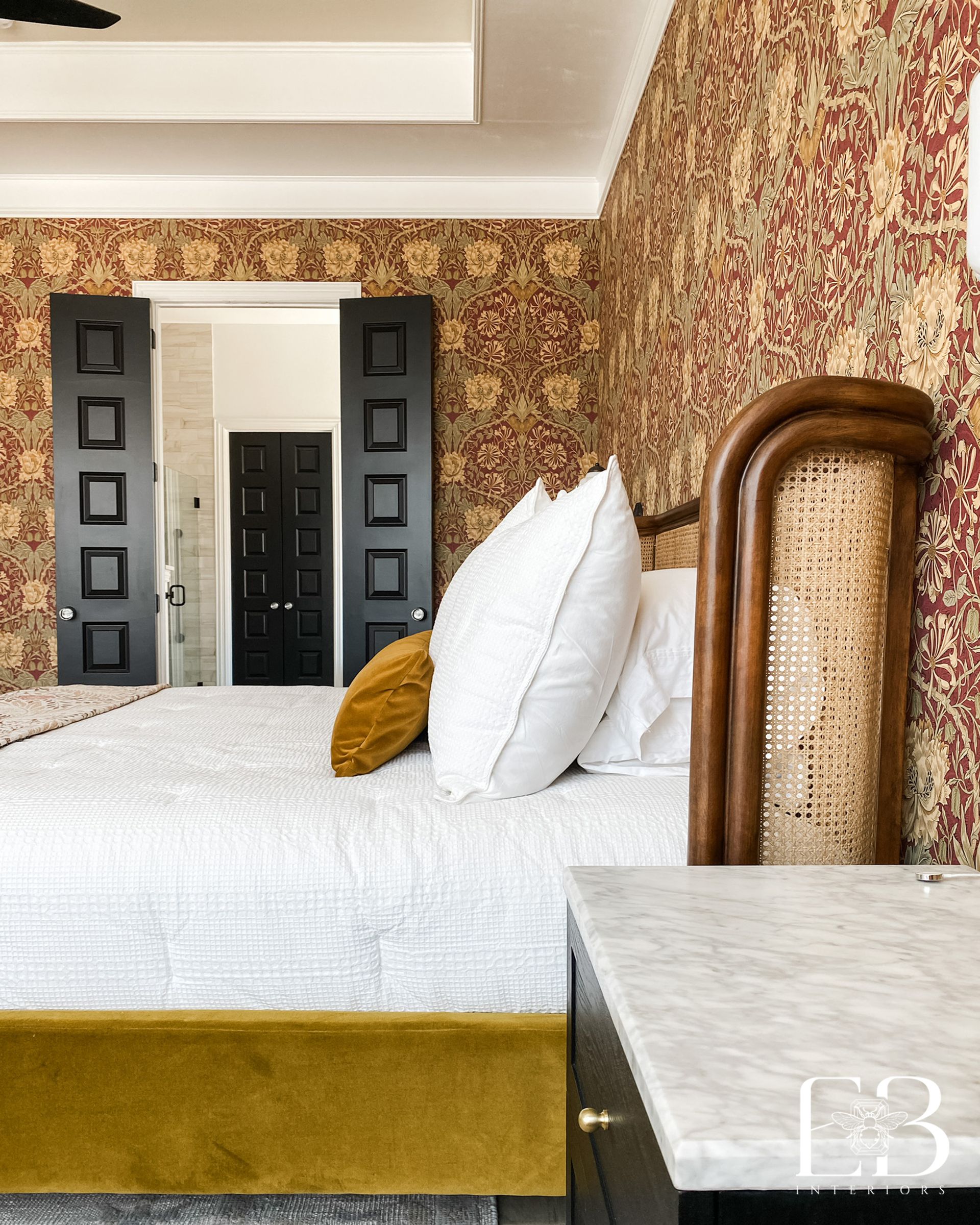 Bedroom with ornate red wallpaper, bed with yellow velvet base, and a headboard. Black doors lead to another room.