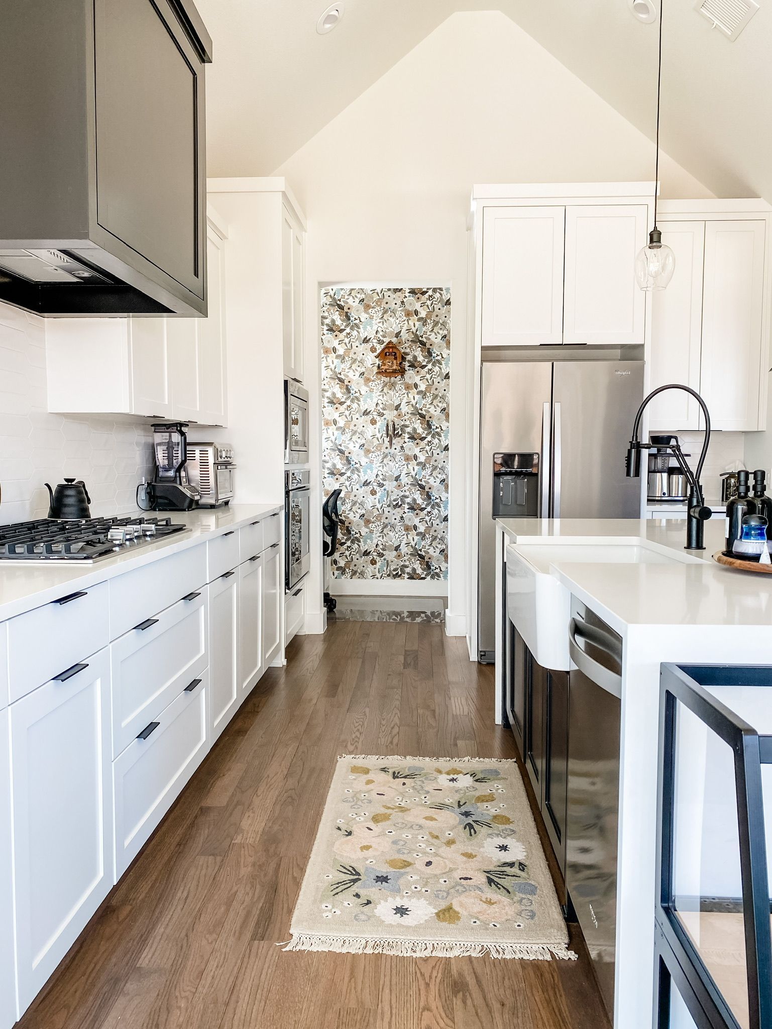 Transitional kitchen design by Emerald Bee Interiors featuring white cabinetry, oak flooring, black accents, and a floral wallpaper nook. Designed by Emily Mussett, Texas-based interior designer.