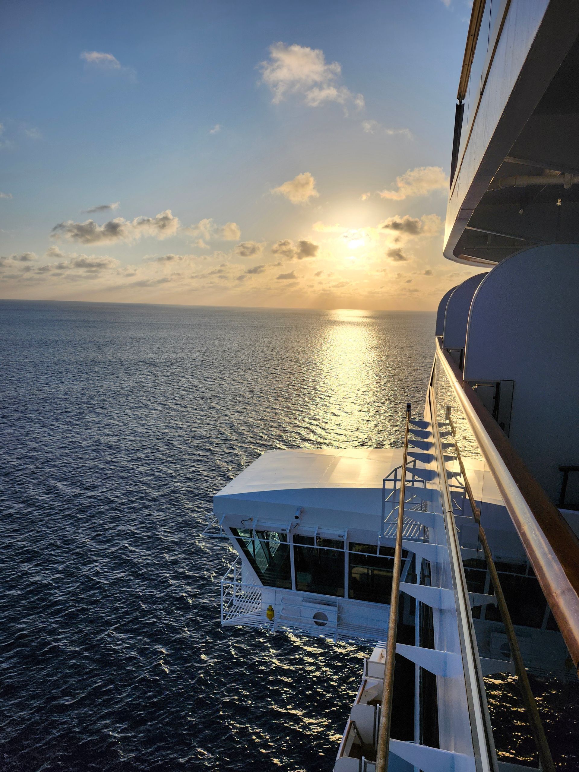 Sunrise over ocean, viewed from cruise ship deck. Sun reflects on water, ship structure visible.