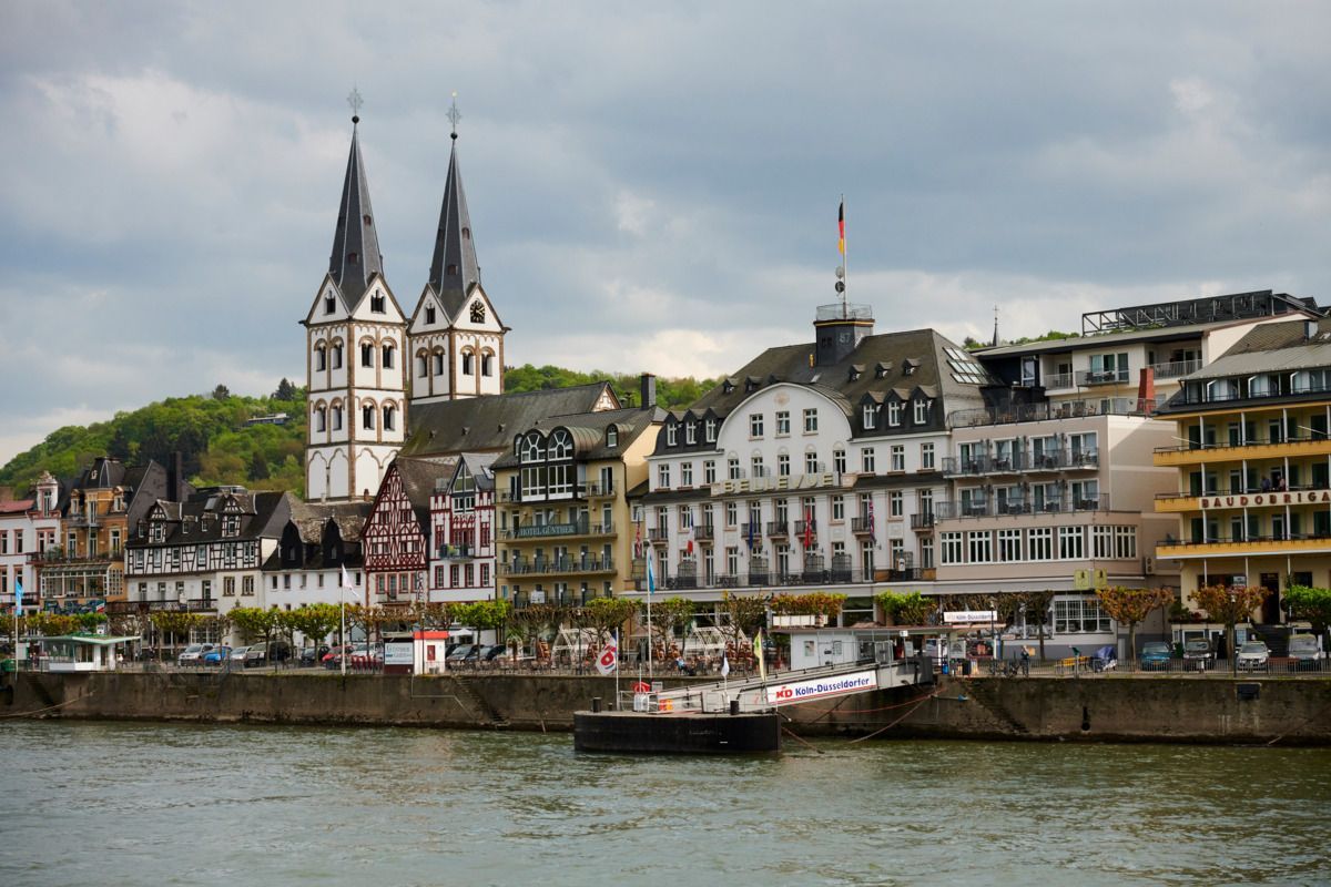 Riverside town with twin church spires, colorful buildings, and boats on a cloudy day