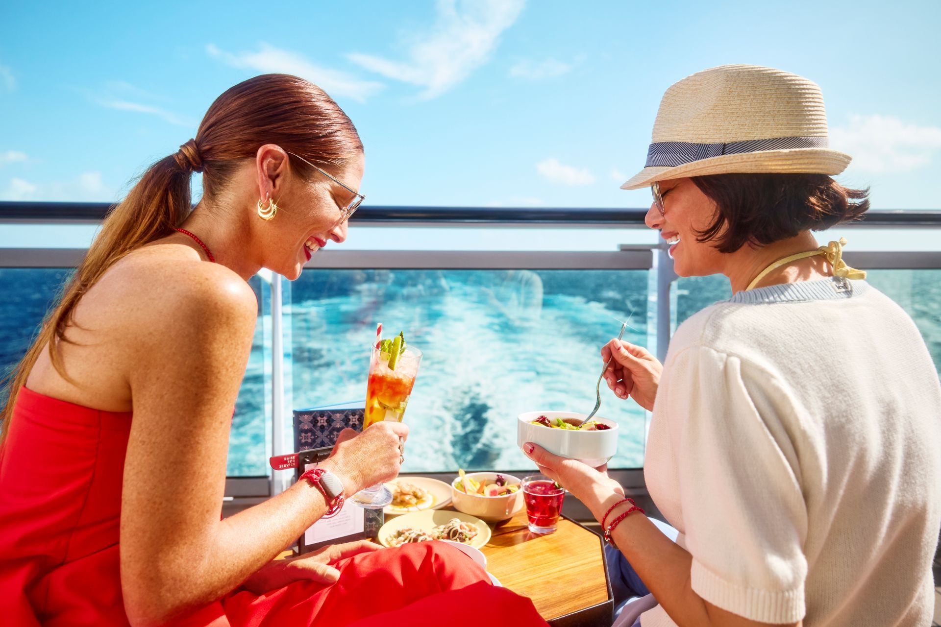 Two people on a cruise ship deck enjoying food and drinks, ocean view in background.