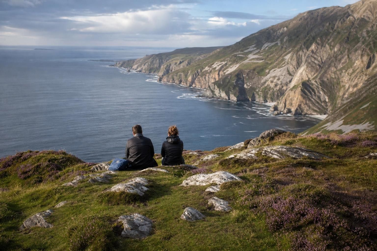 Two people sitting on a grassy cliff overlooking a rocky coastline and calm blue sea