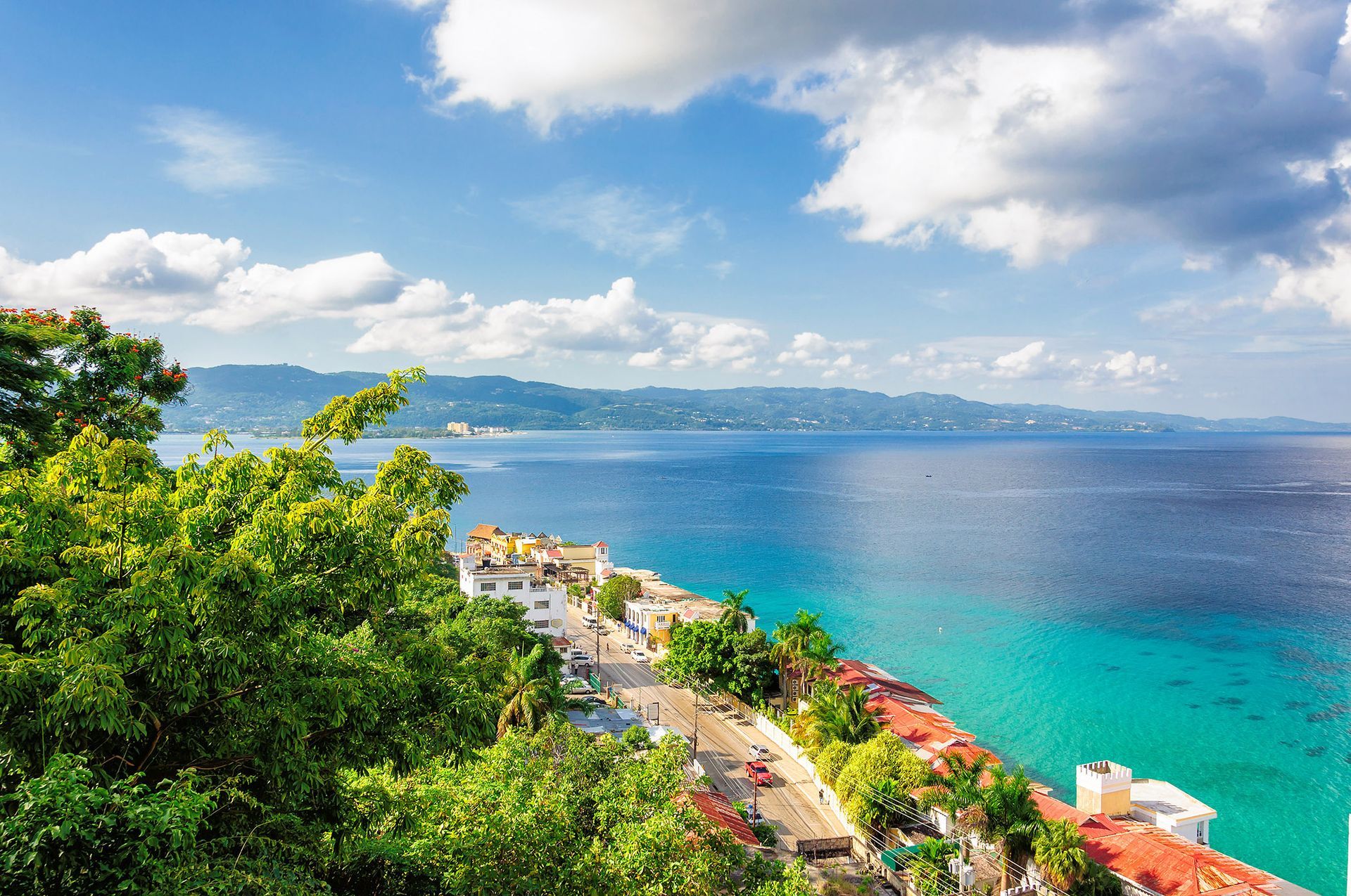 Ocean view with blue water, white buildings, green trees, and a blue sky with clouds.