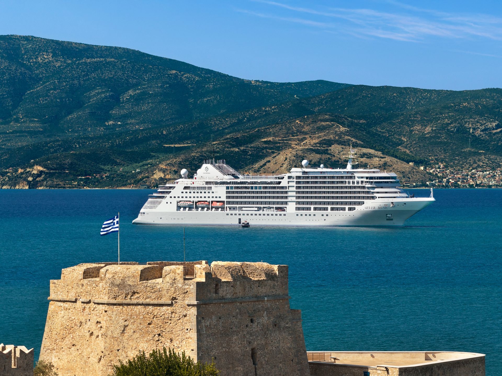 White cruise ship on blue sea near a stone fortress with a Greek flag and mountains in the background