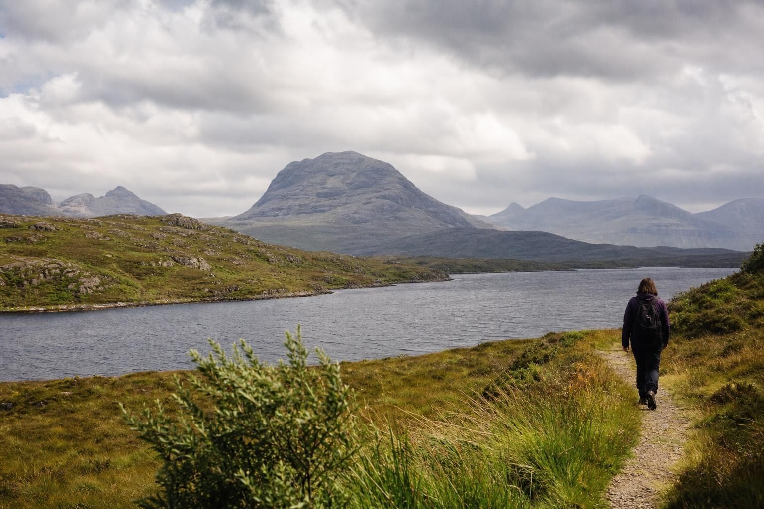 Person walking along a grassy path beside a lake with mountains under cloudy skies