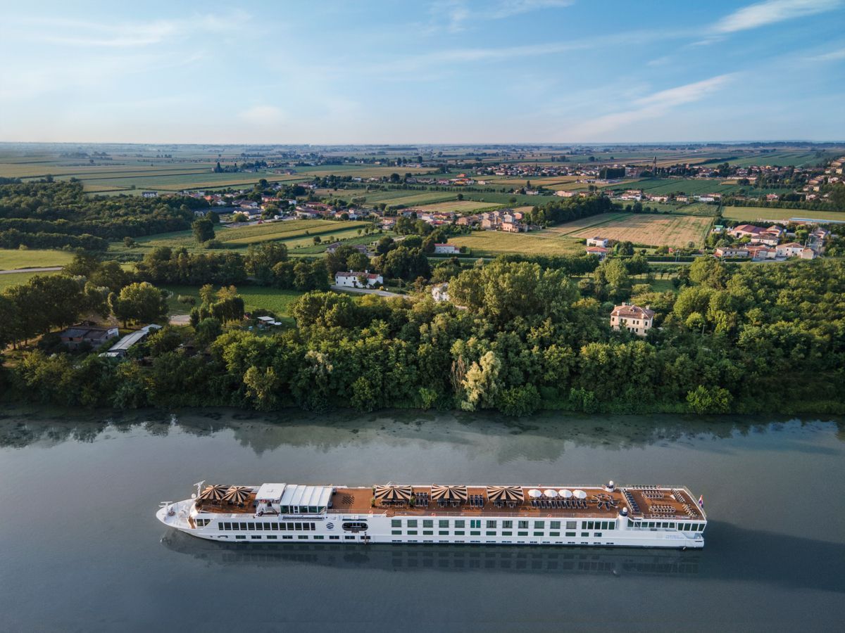River cruise ship on a calm river beside green farmland and a distant village under a clear sky