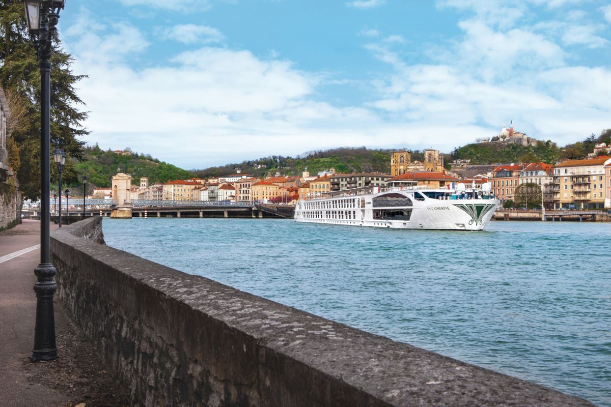 Riverside promenade with stone wall, lampposts, and a ferry crossing a harbor town under cloudy skies