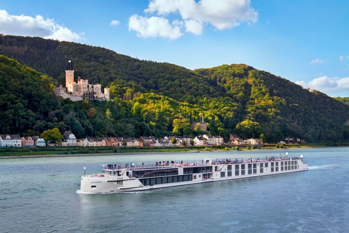 River cruise boat on a wide river with a forested hillside, castle, and village along the shore
