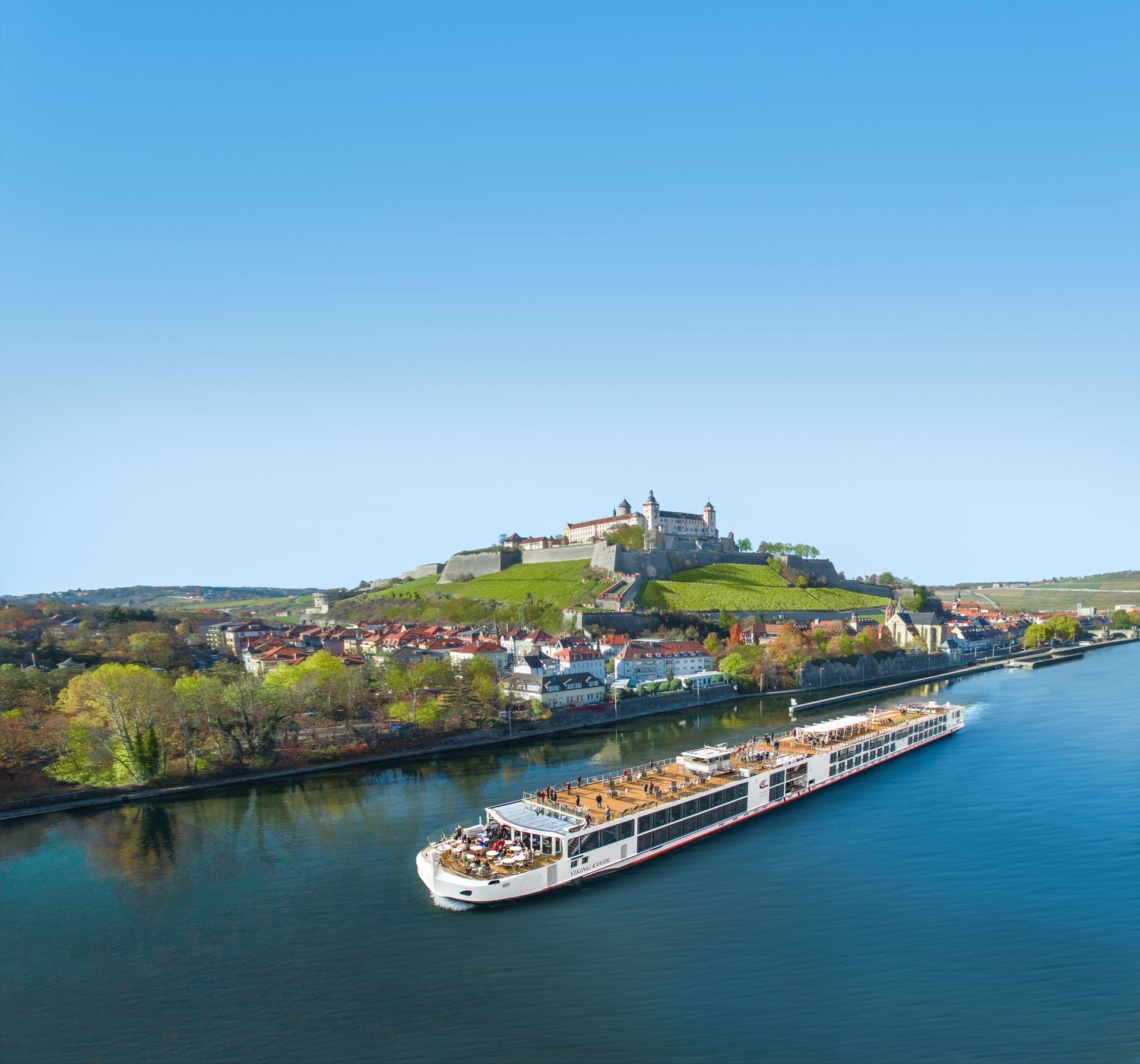 River cruise ship on a blue river beside a hilltop castle and colorful town under a clear sky