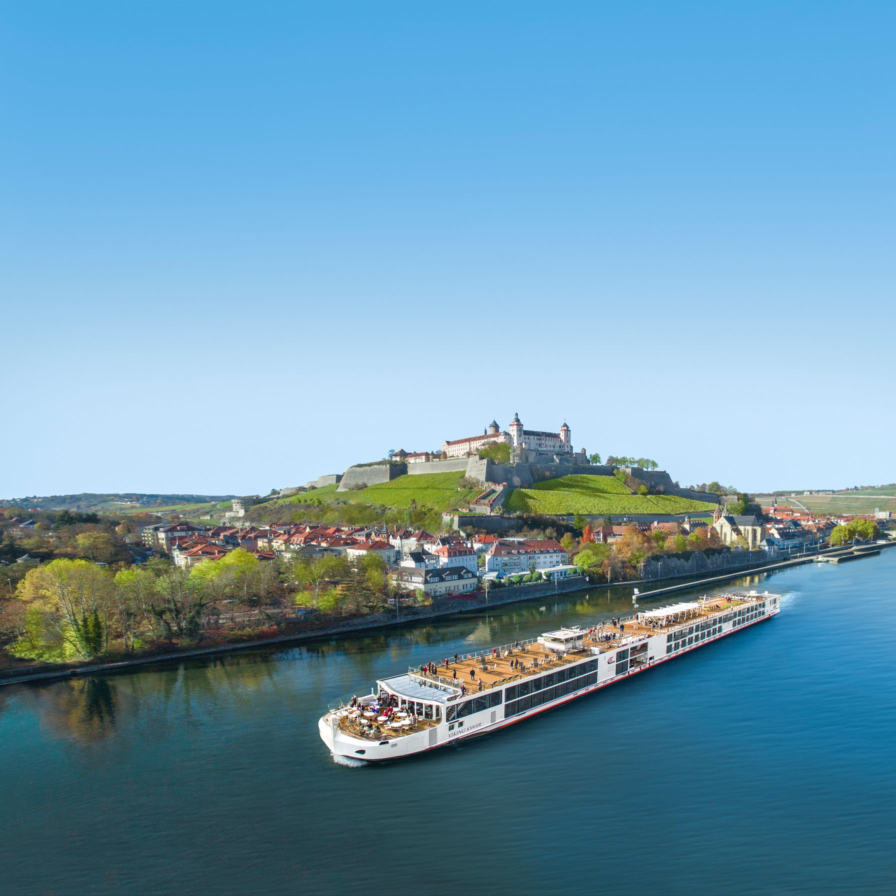 River cruise ship on a blue river beside a hilltop castle and colorful town under a clear sky