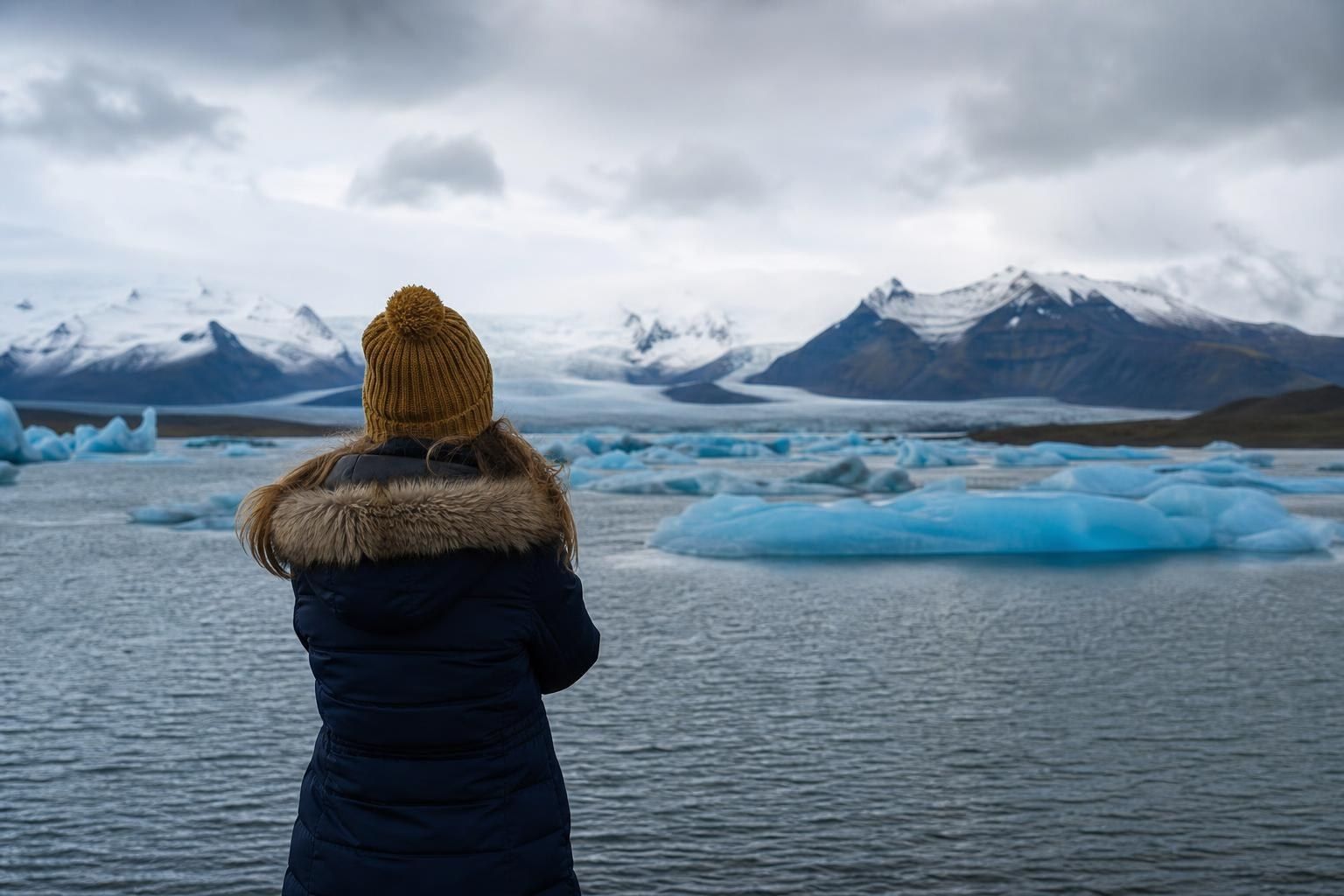 Person in a yellow beanie and dark coat overlooking icy blue icebergs in a snowy fjord
