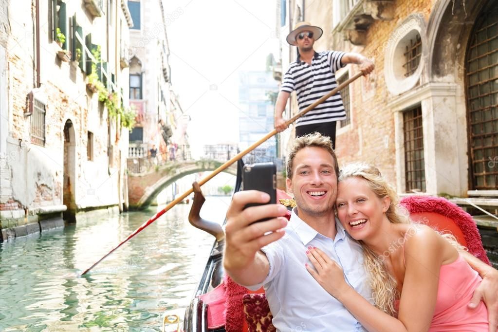 Couple taking a selfie on a gondola in a sunny canal, with colorful buildings beside the water.