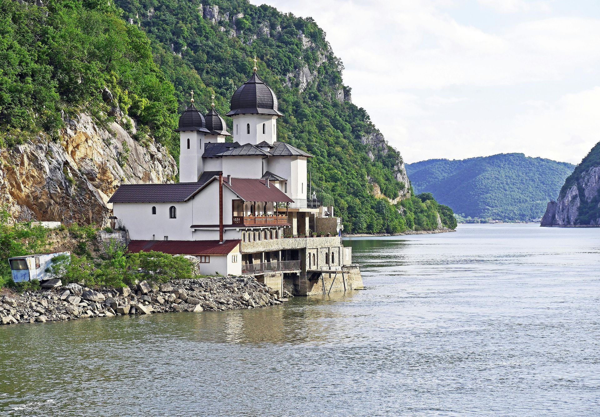 White building with domes on a rocky cliffside, overlooking a wide river. Green trees and hills in background.