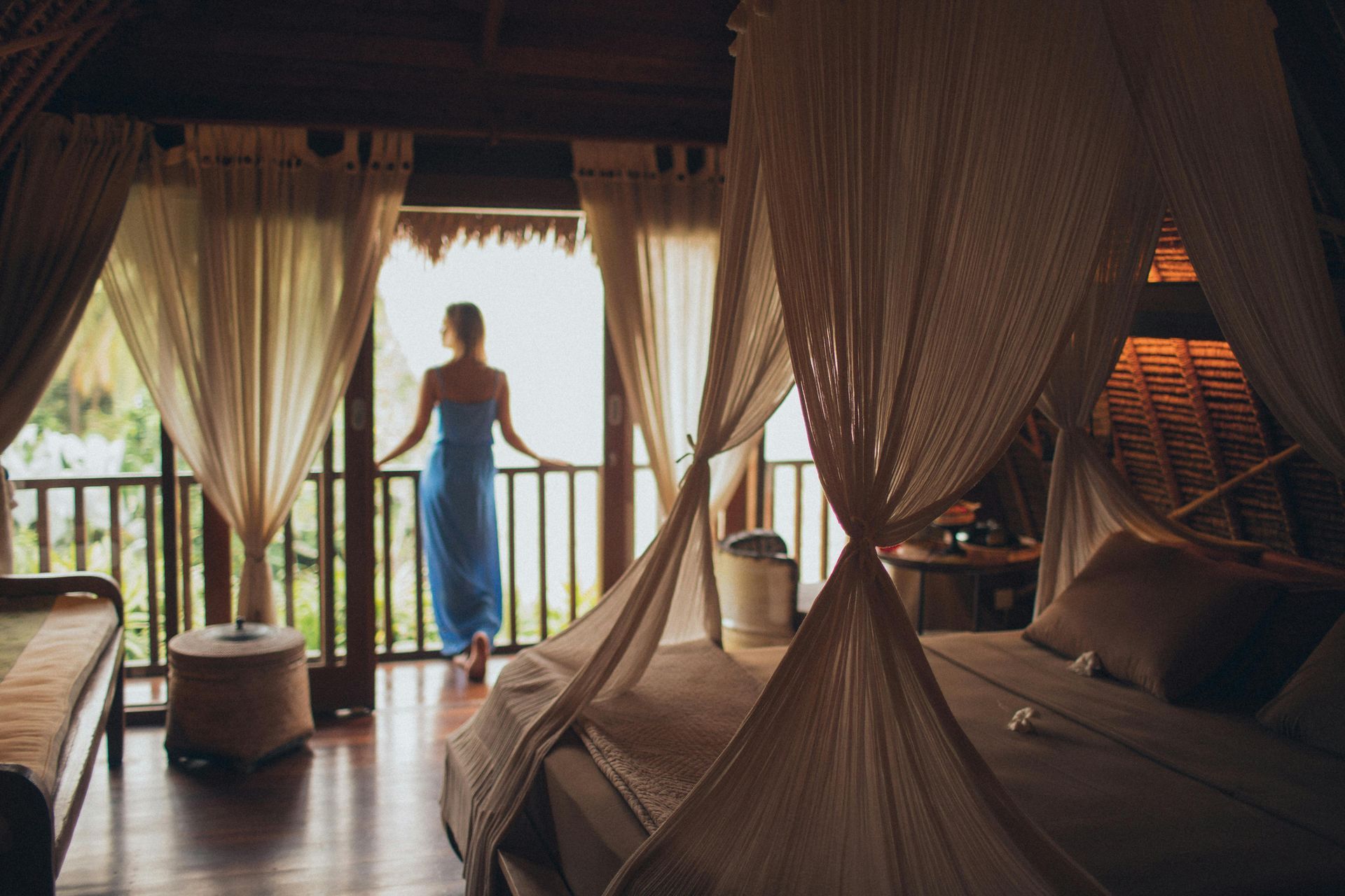 Person standing on a balcony in a tropical, draped bedroom with a canopy bed and ocean view