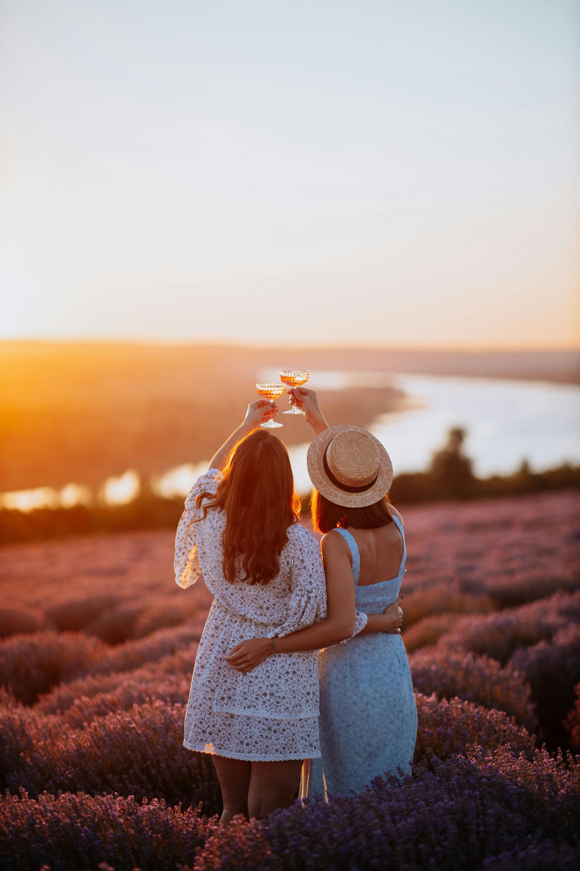 Two people in dresses raise glasses for a toast in a lavender field at sunset.