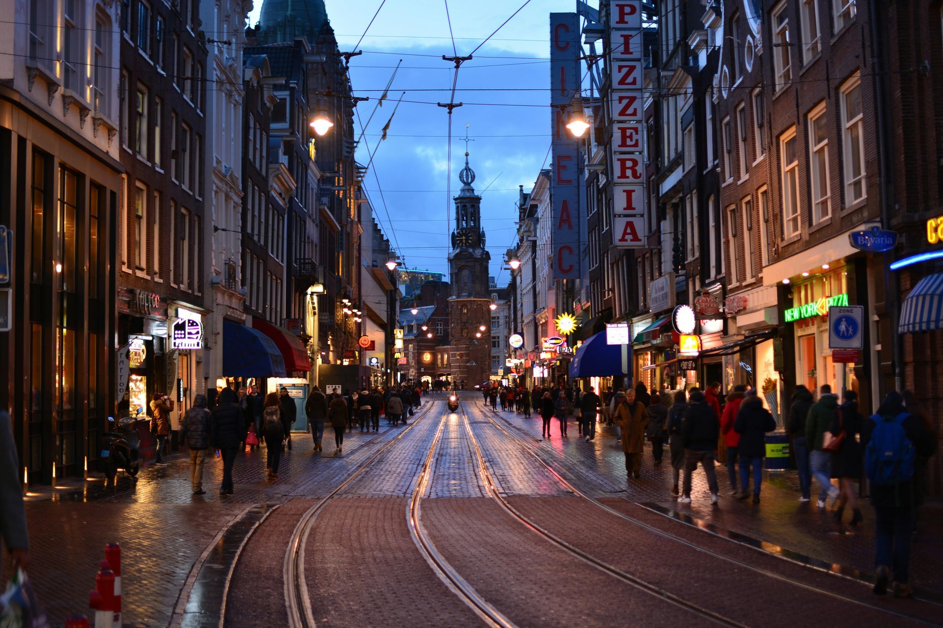 Street scene at dusk in Amsterdam, Netherlands; tram tracks, pedestrians, illuminated buildings, church spire in distance.