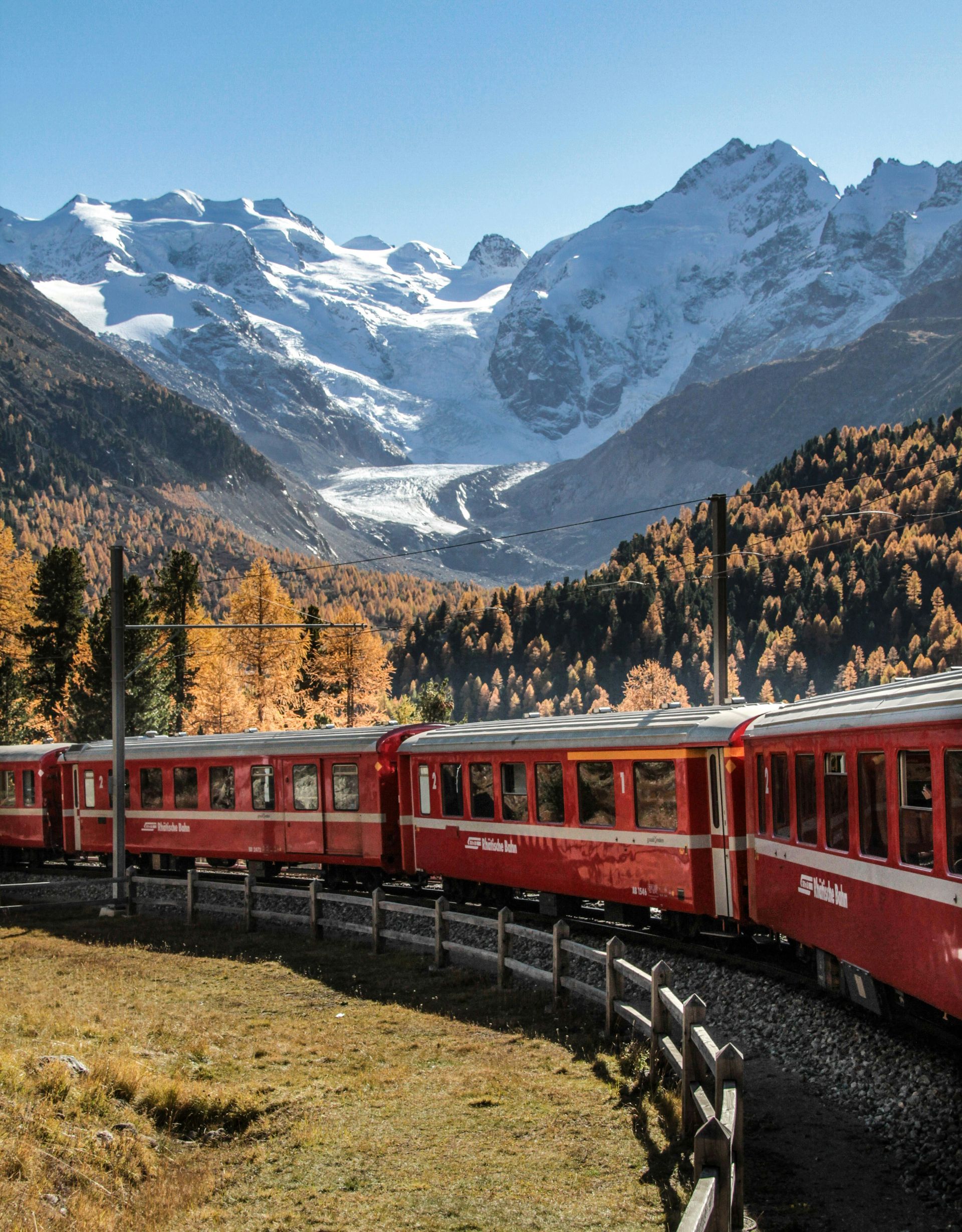 Red train curving through alpine valley with autumn trees and snow-covered mountains beyond