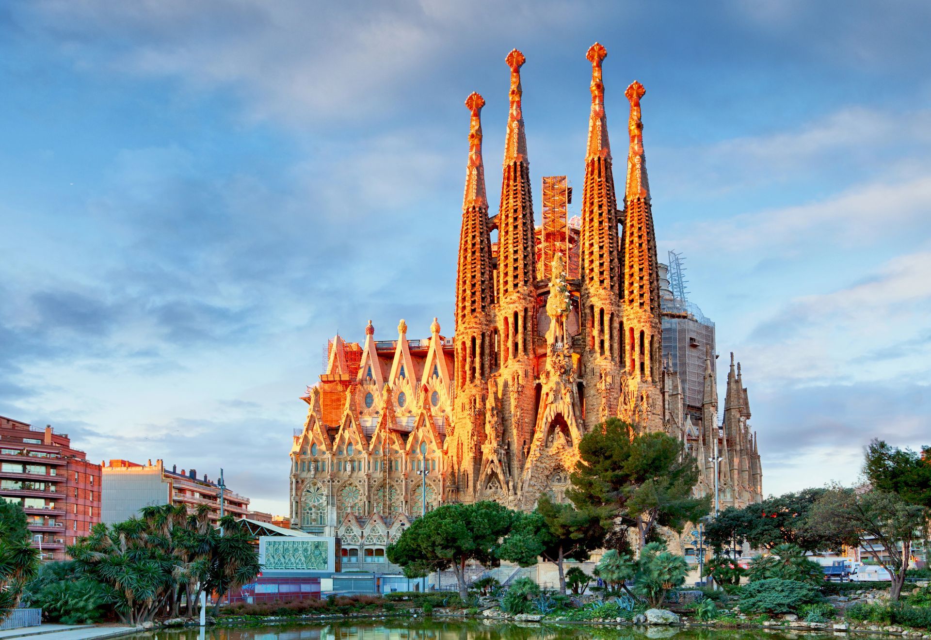 Sagrada Família basilica glowing in warm light under a blue sky, with trees and city buildings in front