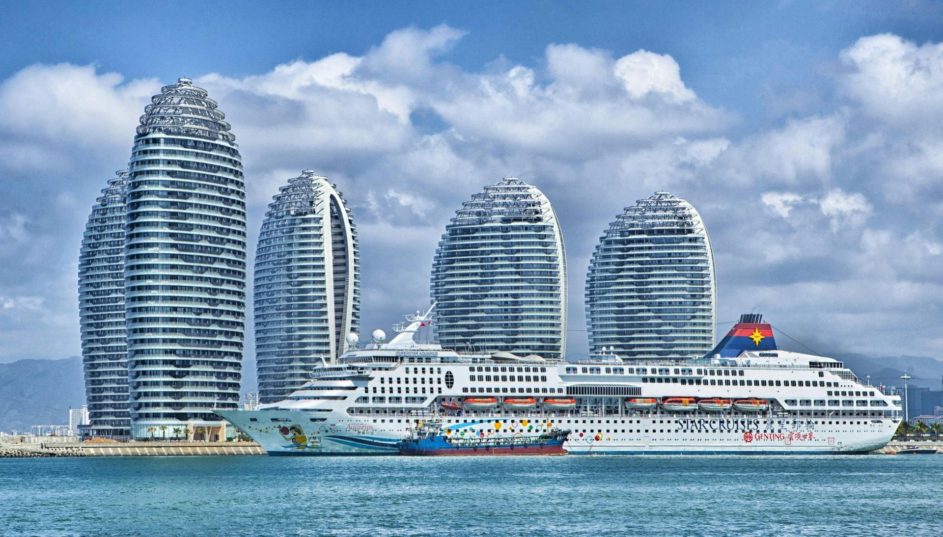 Cruise ship docked in front of five unique, modern buildings along a coastline under a cloudy sky.