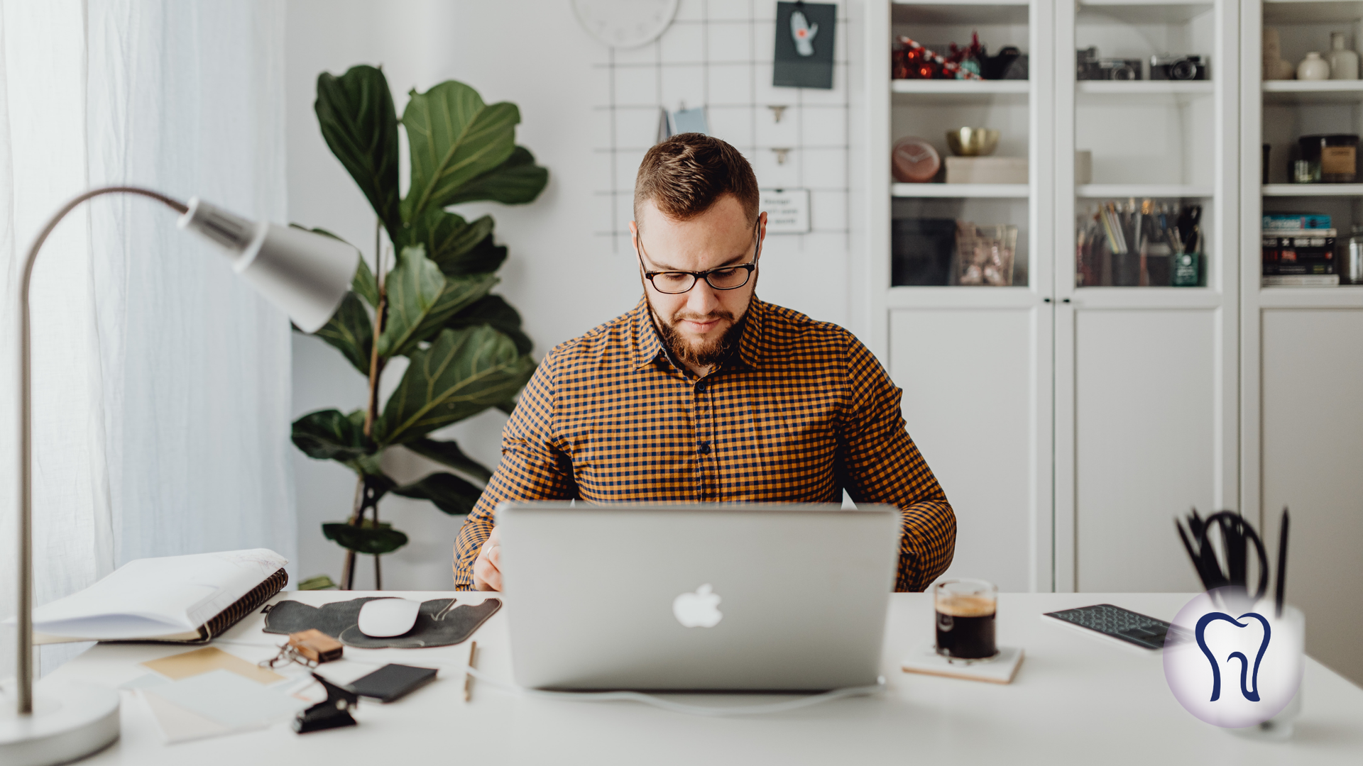 Man working on laptop at a white desk with a plant, lamp, and coffee; shelves in background.