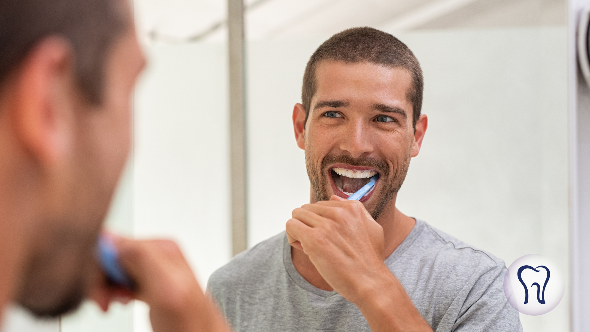 Man brushing his teeth in front of a mirror, smiling, wearing a gray shirt, in a bathroom.