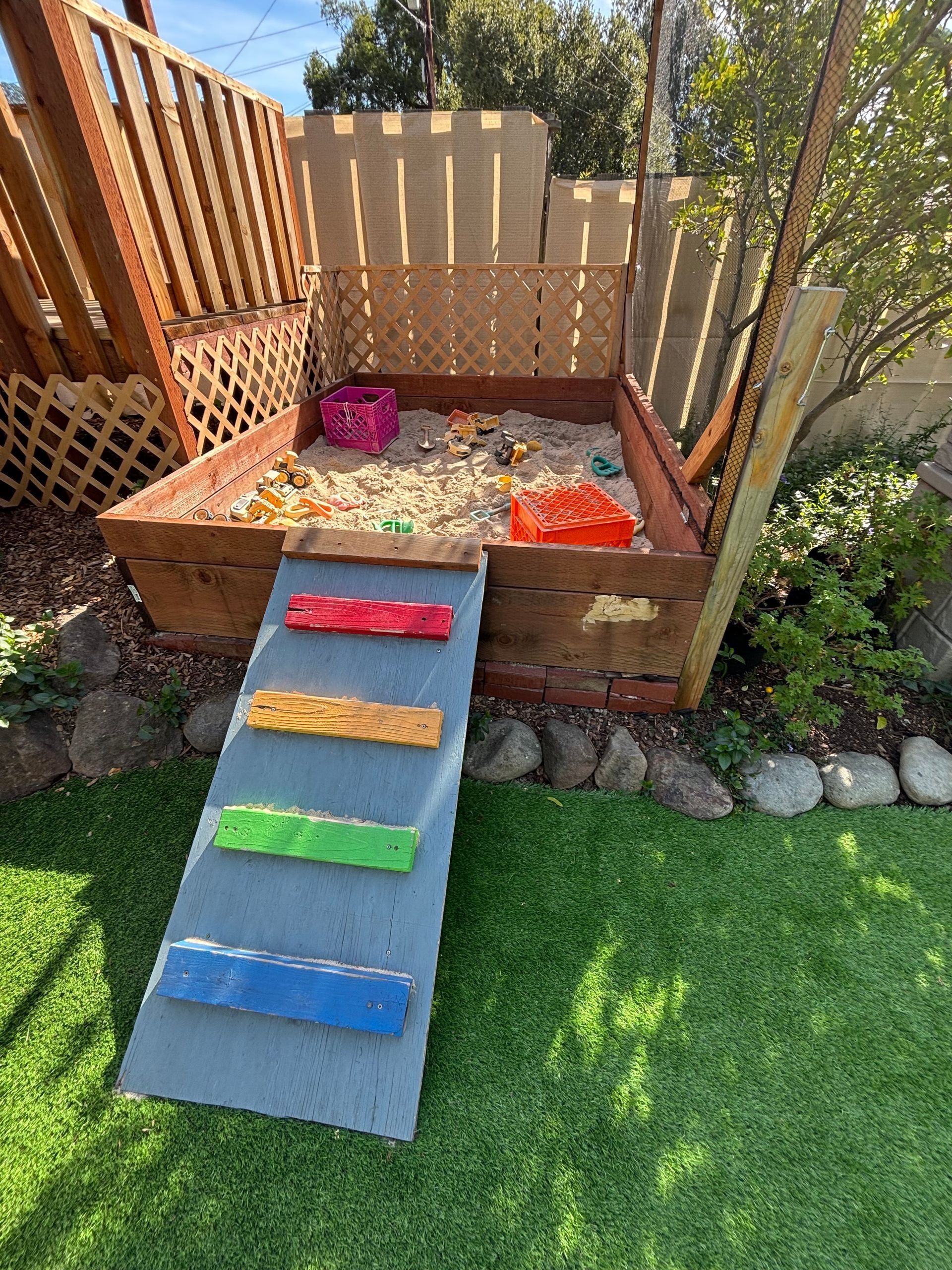 Small wooden playhouse with a ramp painted in rainbow stripes in a sunny garden.