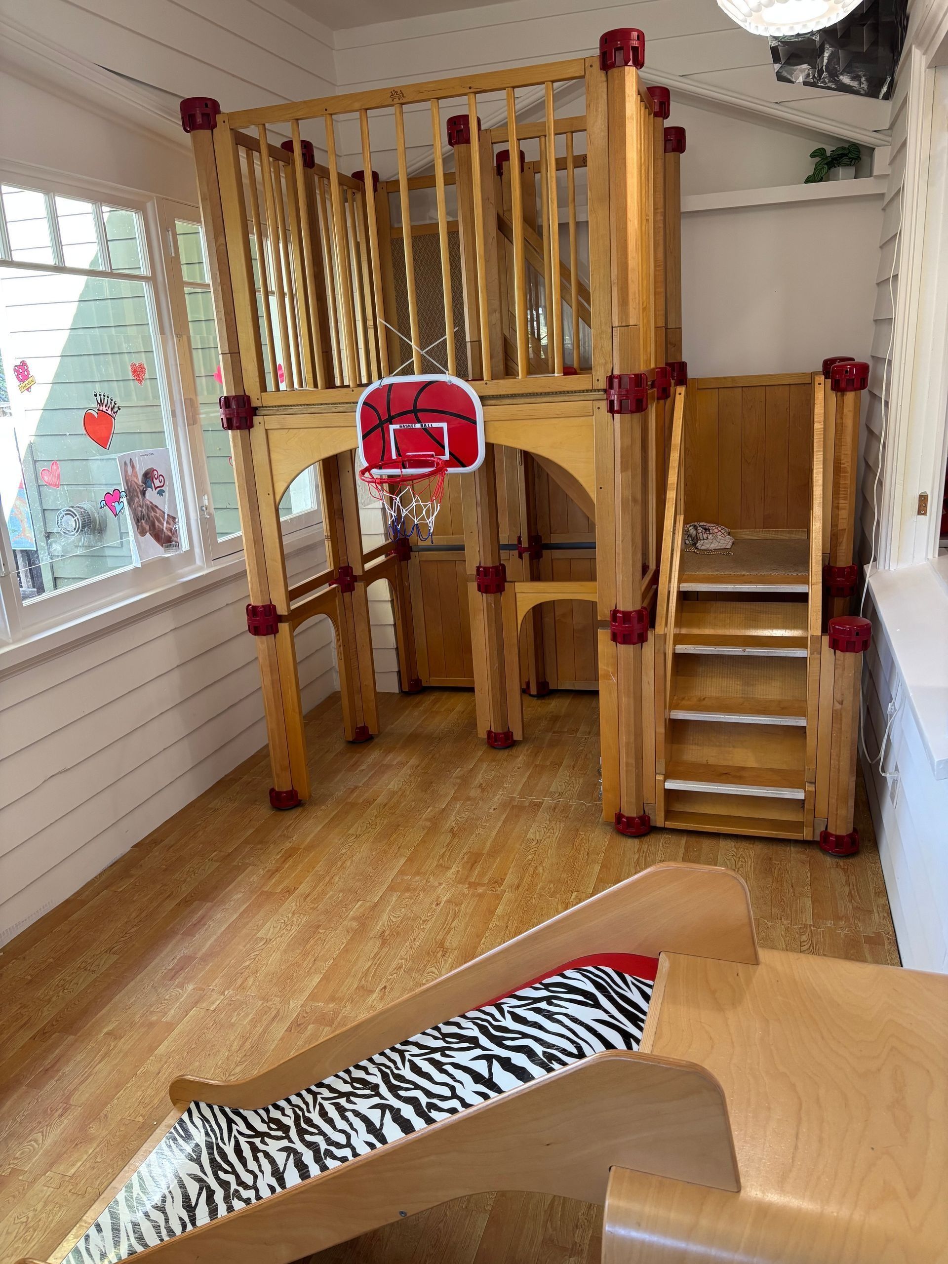 Indoor wooden play structure with ladder, slide, and red basketball hoop in a playroom.