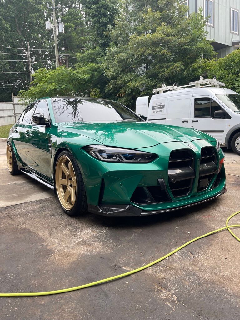 A green BMW M3 is parked in a driveway next to a white van. We provided the wash and wax, exterior Detailing, and Ceramic Coating. 